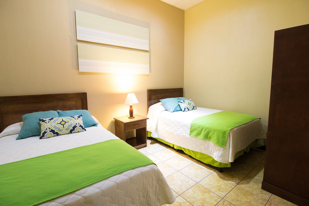 Habitación de hotel con dos camas, cada una con cubrecama verde y almohadas a juego, una mesita de noche con lámpara y una ventana con persianas.