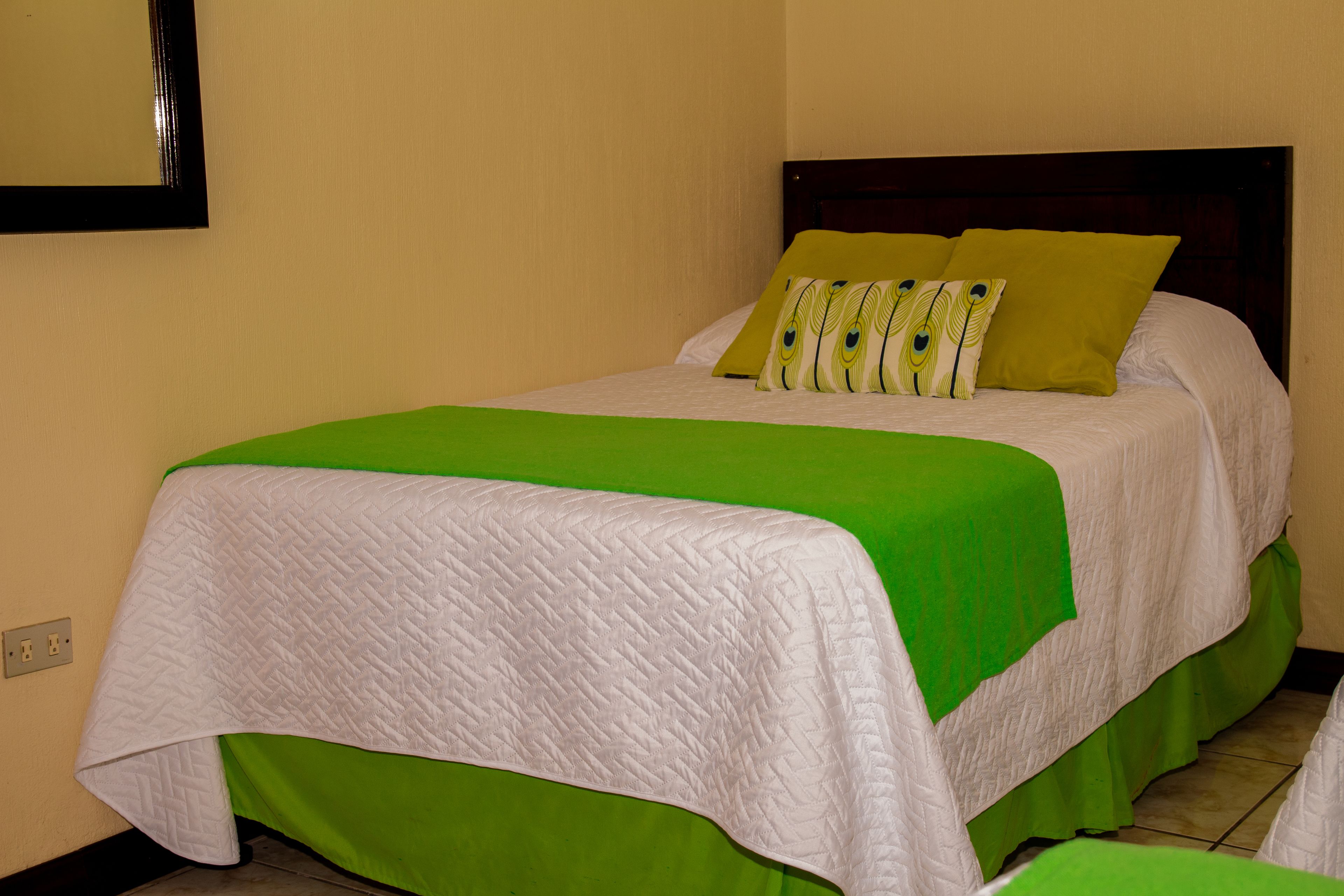A bed with a green bedspread and white quilted cover, two pillows with a leaf pattern, and a dark wooden headboard.