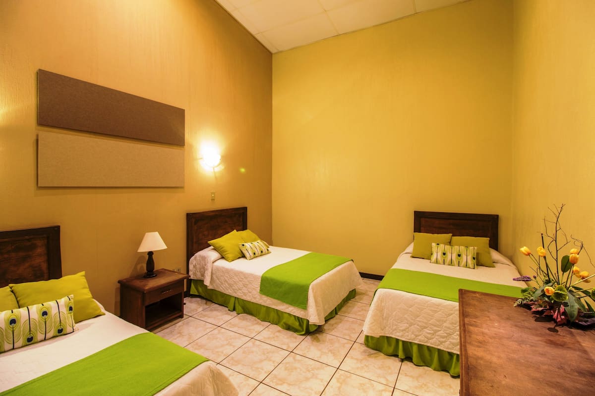Habitación de hotel con dos camas, cada una con ropa de cama verde, una mesita de noche con lámpara y un cabecero de madera.