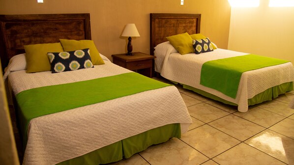 Dos camas con ropa de cama verde y blanca, una mesita de noche con lámpara, y un piso de baldosas.