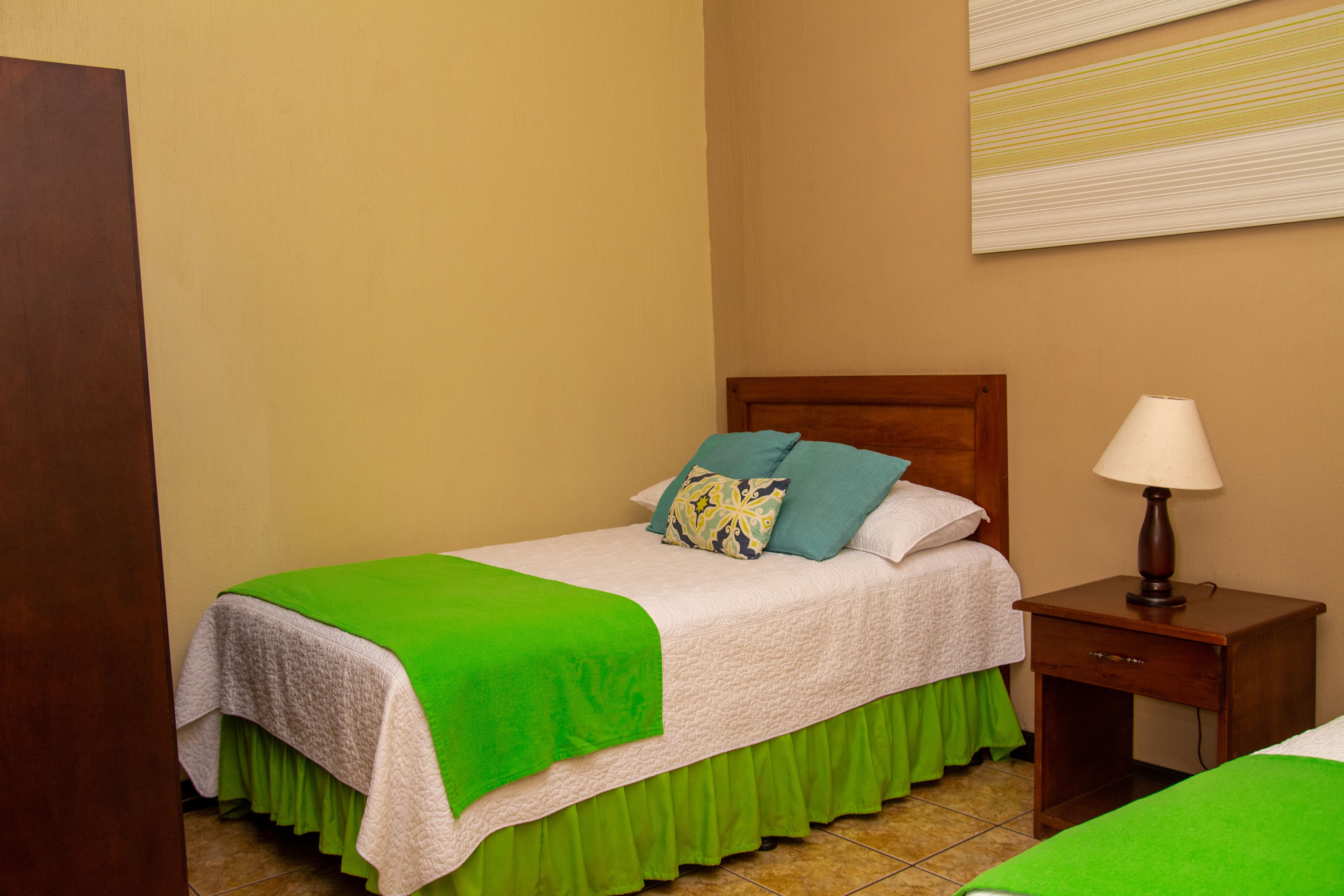 Una cama individual con una colcha verde y ropa de cama blanca, una mesita de noche de madera con una lámpara y una pared con estampado a rayas.