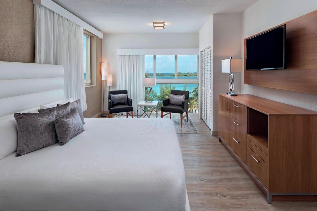 A hotel room with a large bed, a TV mounted on the wall, a desk with two chairs, and a view of the ocean.