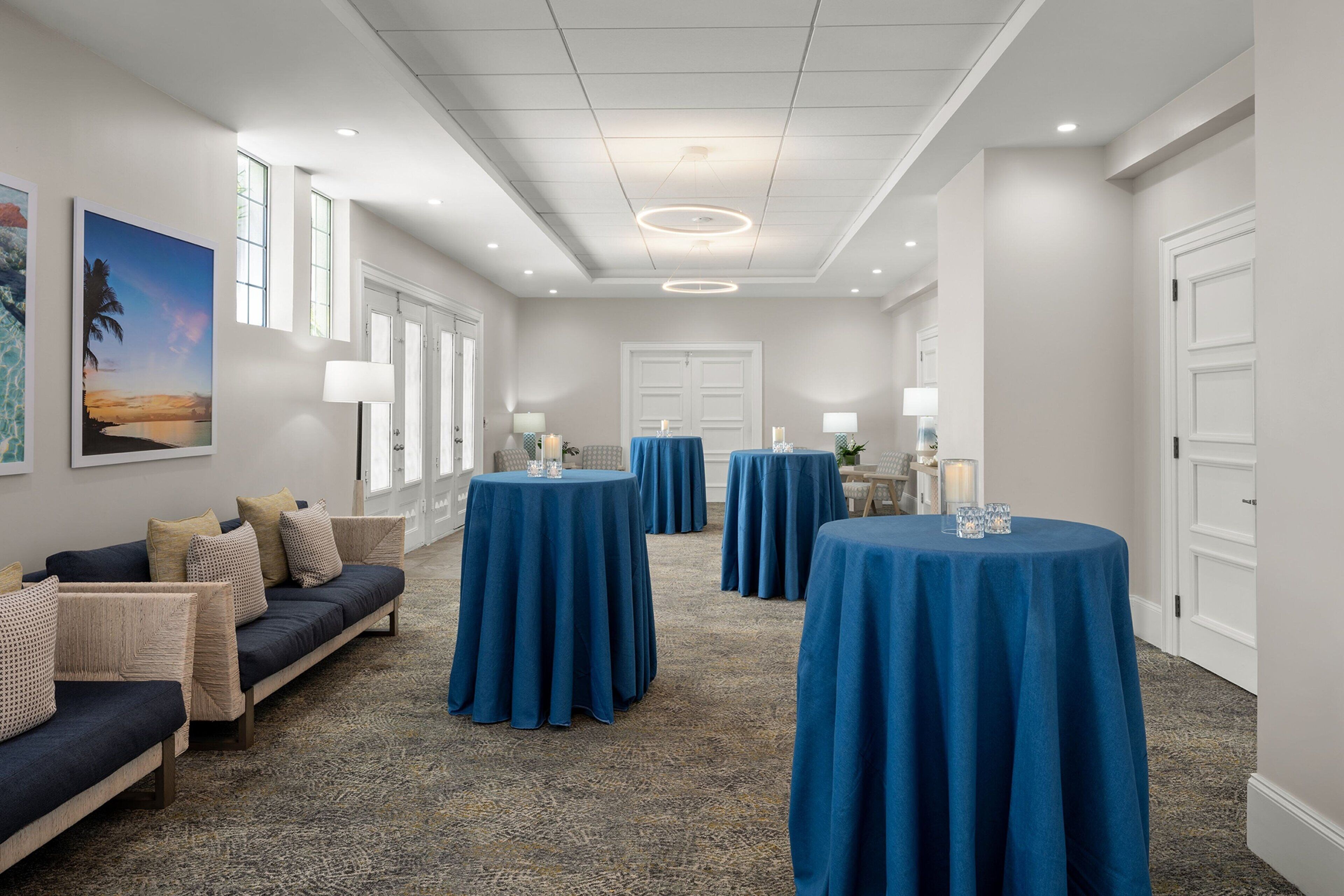 Un hall d’hôtel avec un tapis bleu, un canapé avec des coussins et des tables rondes recouvertes de nappes bleues.