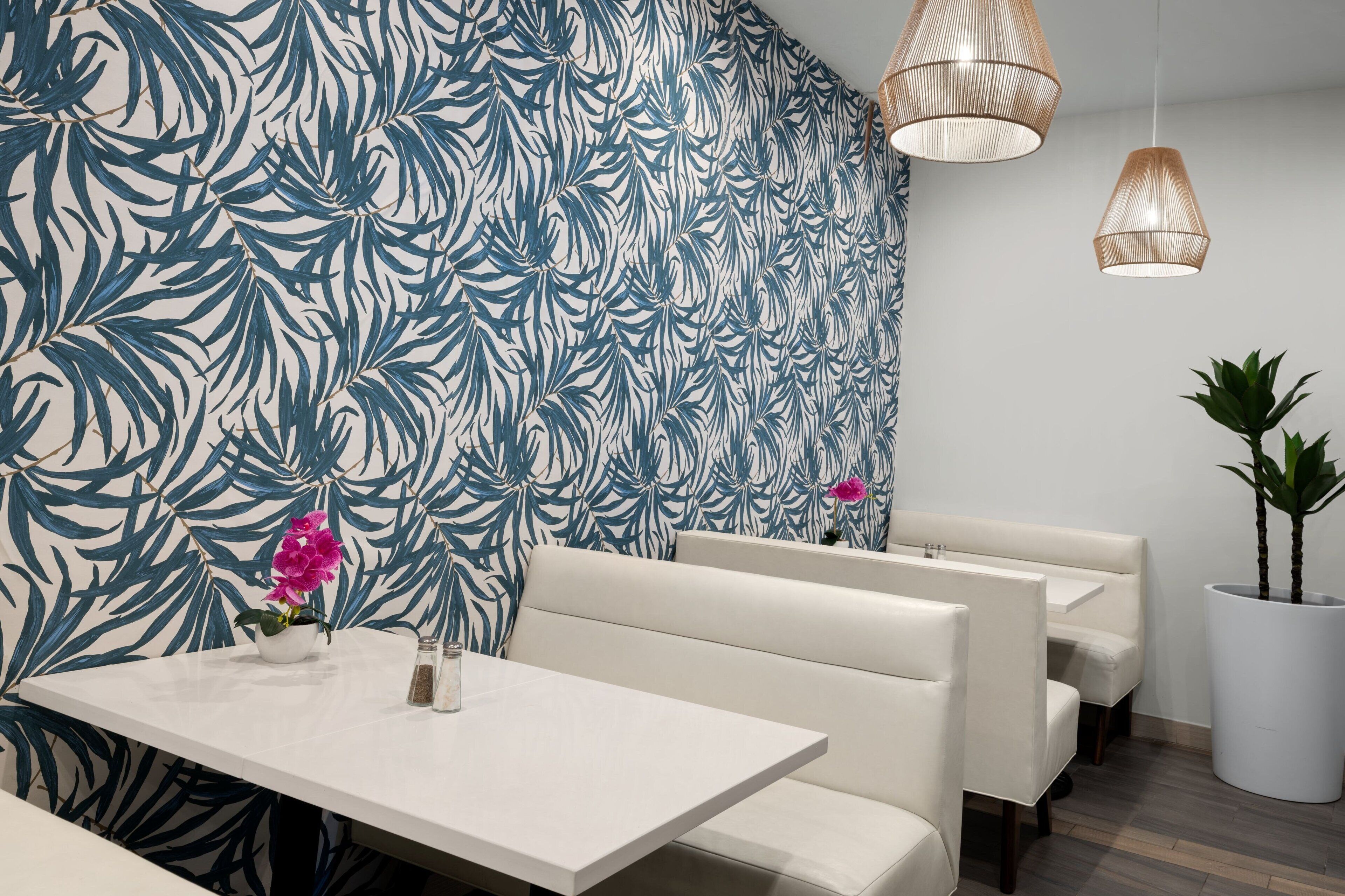 Un restaurante moderno con papel tapiz con estampado de hojas tropicales, asientos tipo cabina acolchados en blanco y lámparas colgantes de mimbre.