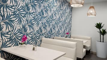 Un restaurante moderno con papel tapiz con estampado de hojas tropicales, asientos tipo cabina acolchados en blanco y lámparas colgantes de mimbre.