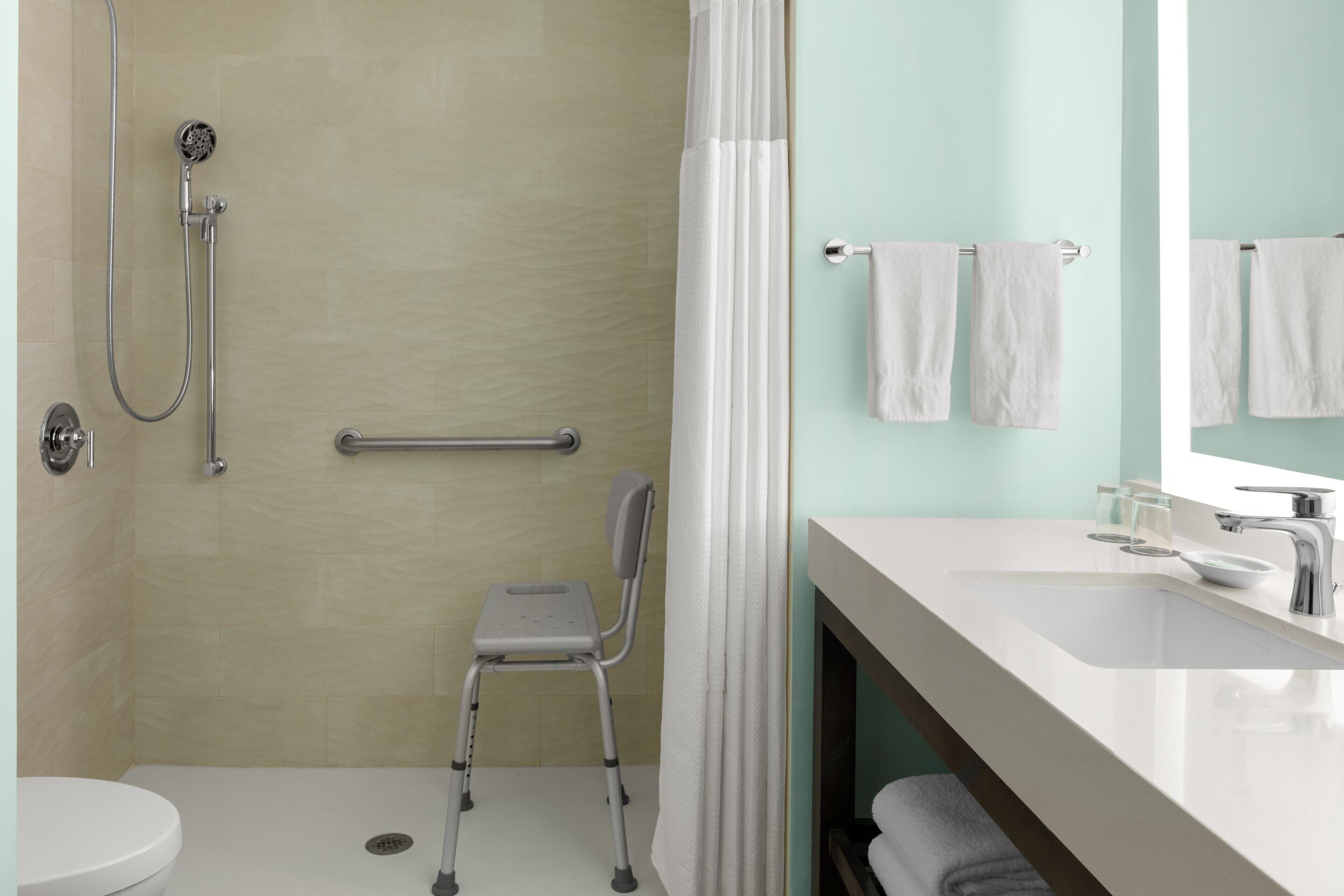 A bathroom with a shower, a toilet, a grab bar, a wheelchair-accessible chair, and a sink with a mirror.