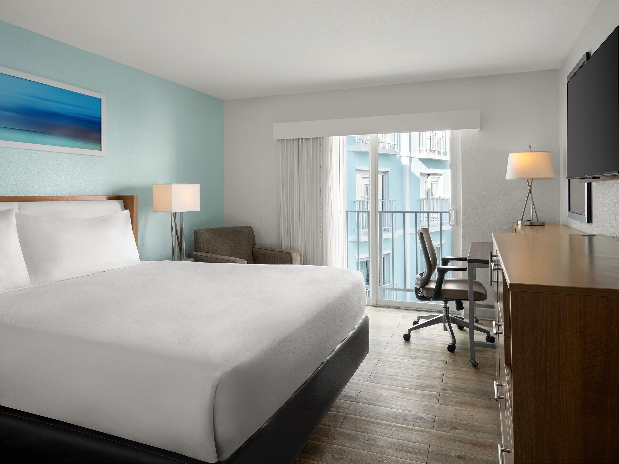 Habitación de hotel con una cama grande, un escritorio, dos sillas, un televisor y un balcón con vistas a edificios.