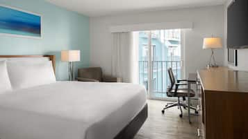 Habitación de hotel con una cama grande, un escritorio, dos sillas, un televisor y un balcón con vistas a edificios.