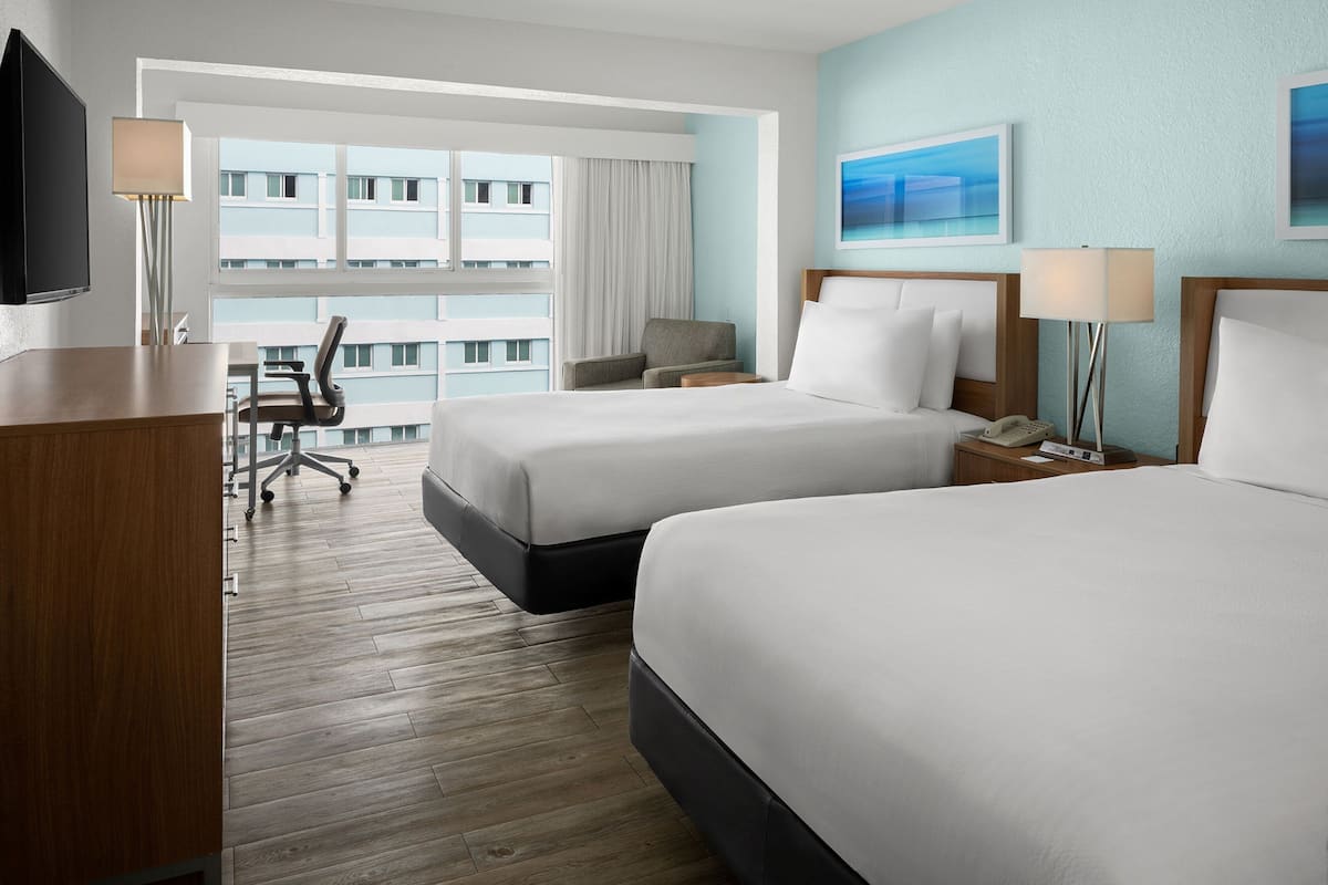 A hotel room with two beds, a desk, a chair, a TV, and a window with a view of a building.