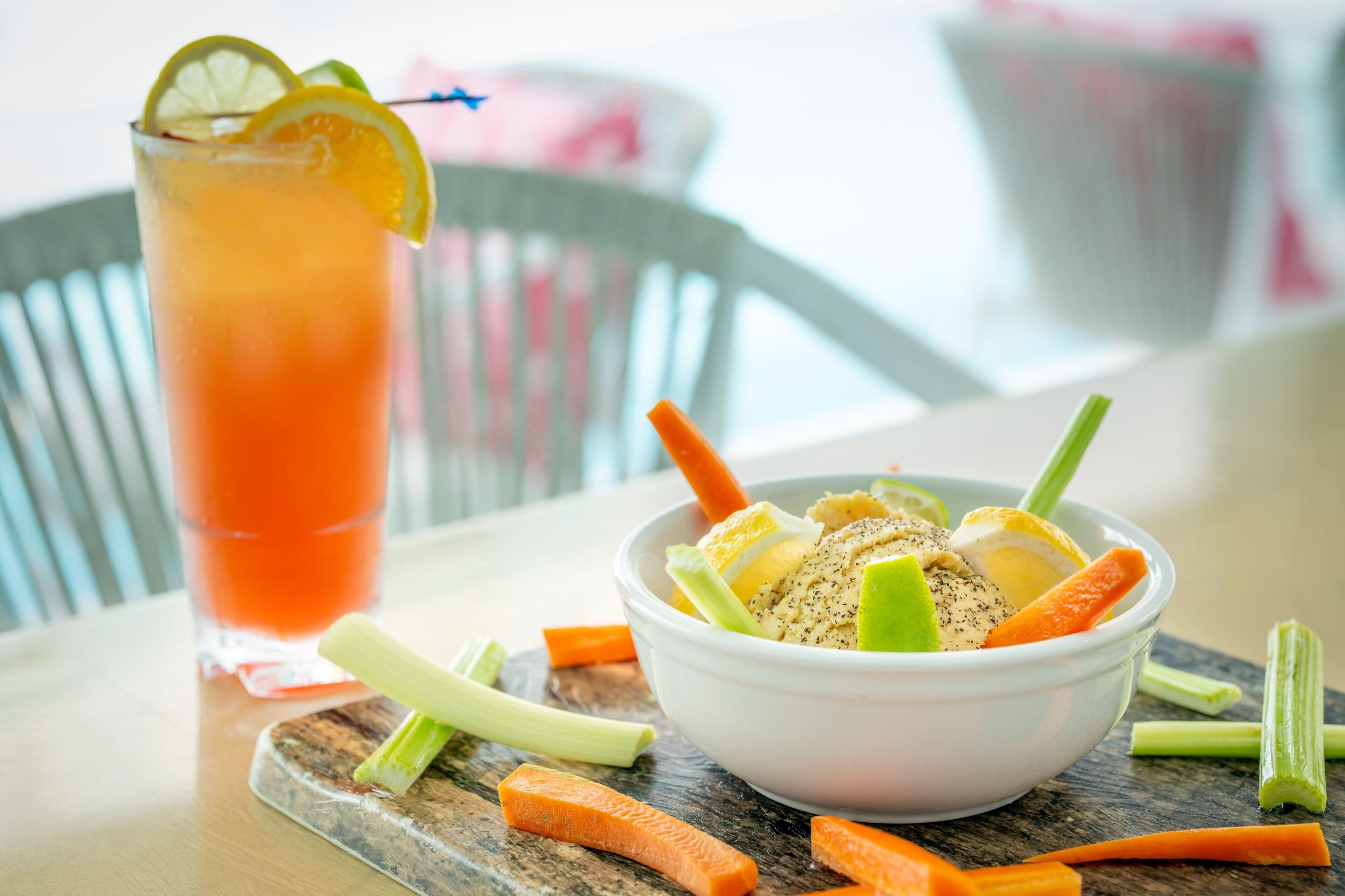 Una mesa con un vaso de jugo de naranja, un tazón de ensalada con apio y zanahorias, y una tabla de cortar de madera.