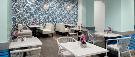 En modern restaurang med vita bord och flätade stolar, med en vägg prydd av ett tropiskt bladmotiv och hängande lampor.