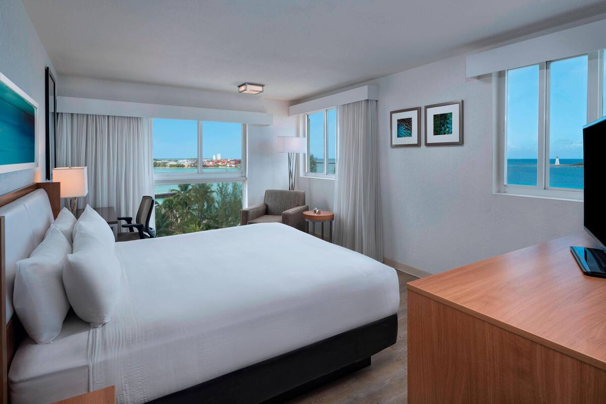 A hotel room with a large bed, a desk, a chair, a lamp, and a view of the ocean.