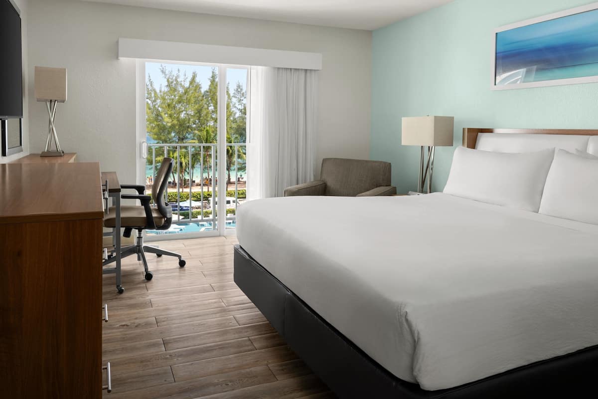 A hotel room with a large bed, a desk with a lamp, a chair, and a view of a pool and trees.