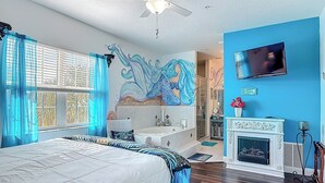 A bedroom with a bed, a ceiling fan, a large window with curtains, a bathroom with a bathtub, and a fireplace. - Inn On the Avenue (New Smyrna Beach)