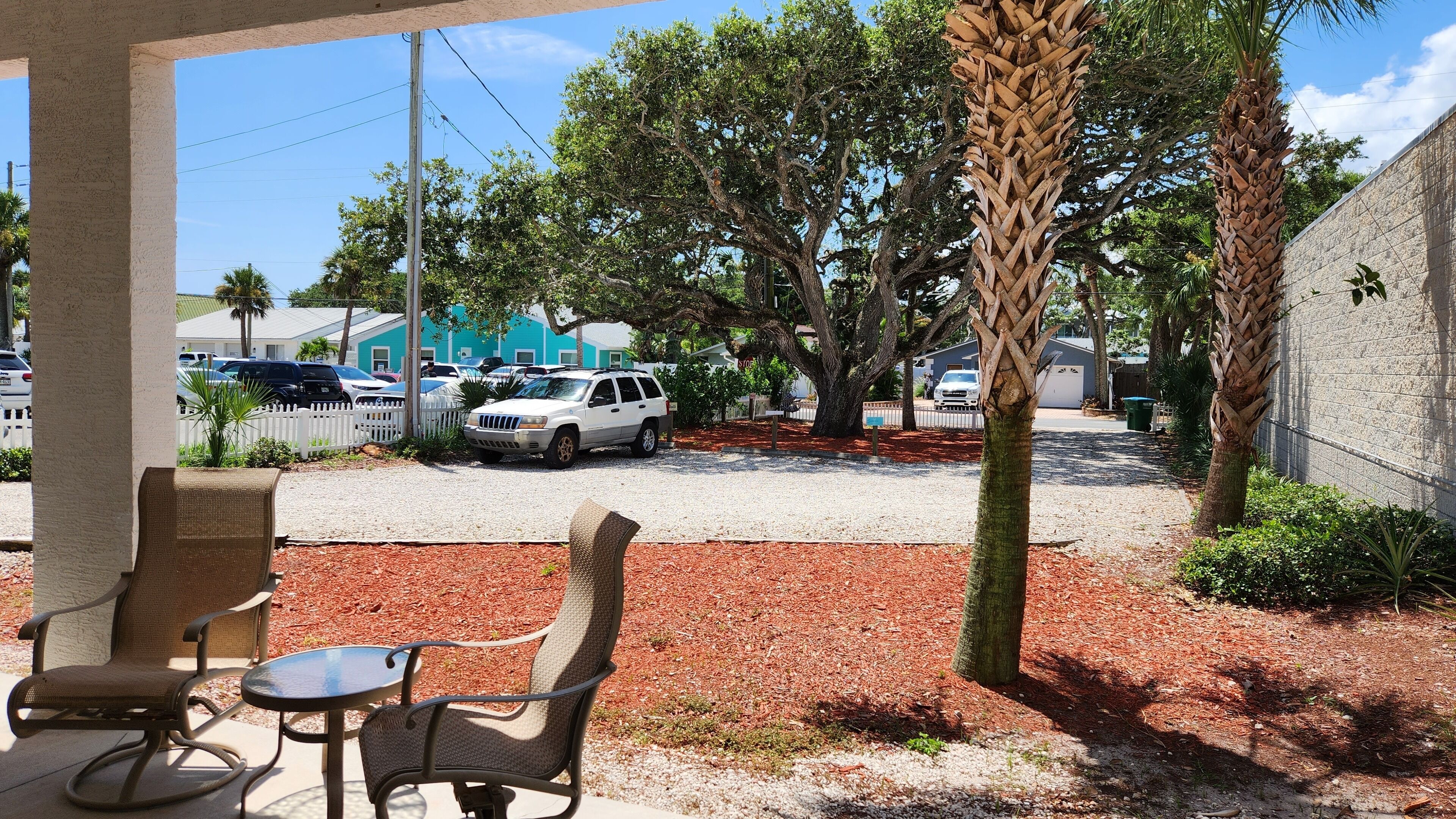 A covered outdoor seating area with wicker chairs and a small table, surrounded by trees and a gravel driveway.