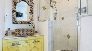 Un baño con tocador amarillo, un lavabo redondo con diseño de mosaico, un espejo decorativo y una ducha con azulejos de patrón de diamante.