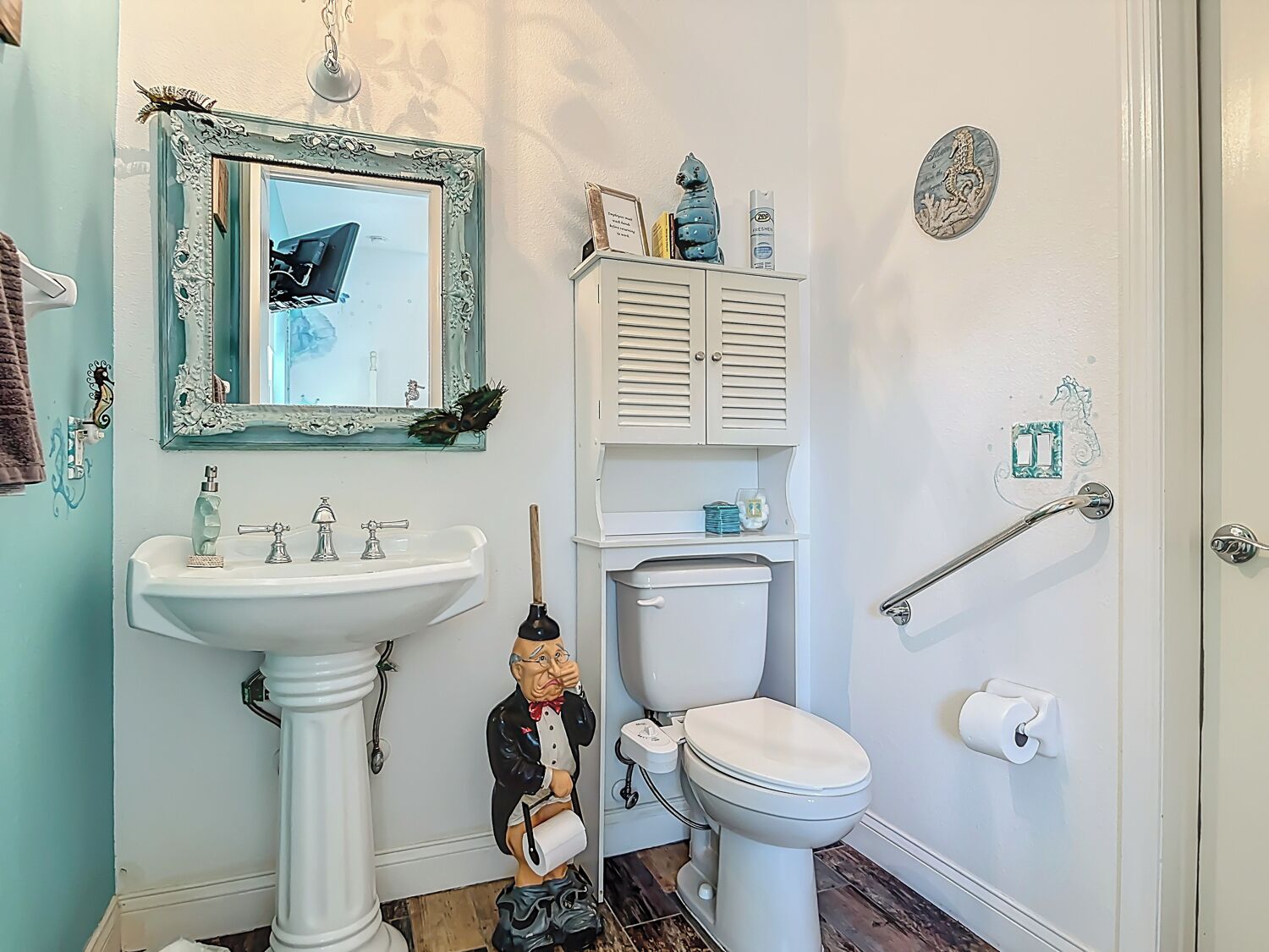 A bathroom with a pedestal sink, a toilet, a mirror with a decorative frame, and a small cabinet with toiletries.