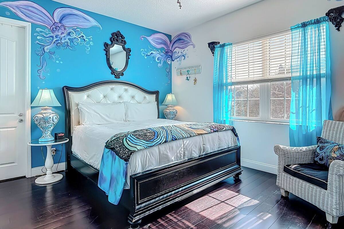 Una camera da letto con un letto grande, una sedia, una finestra con tende e un muro decorato con un affresco floreale.