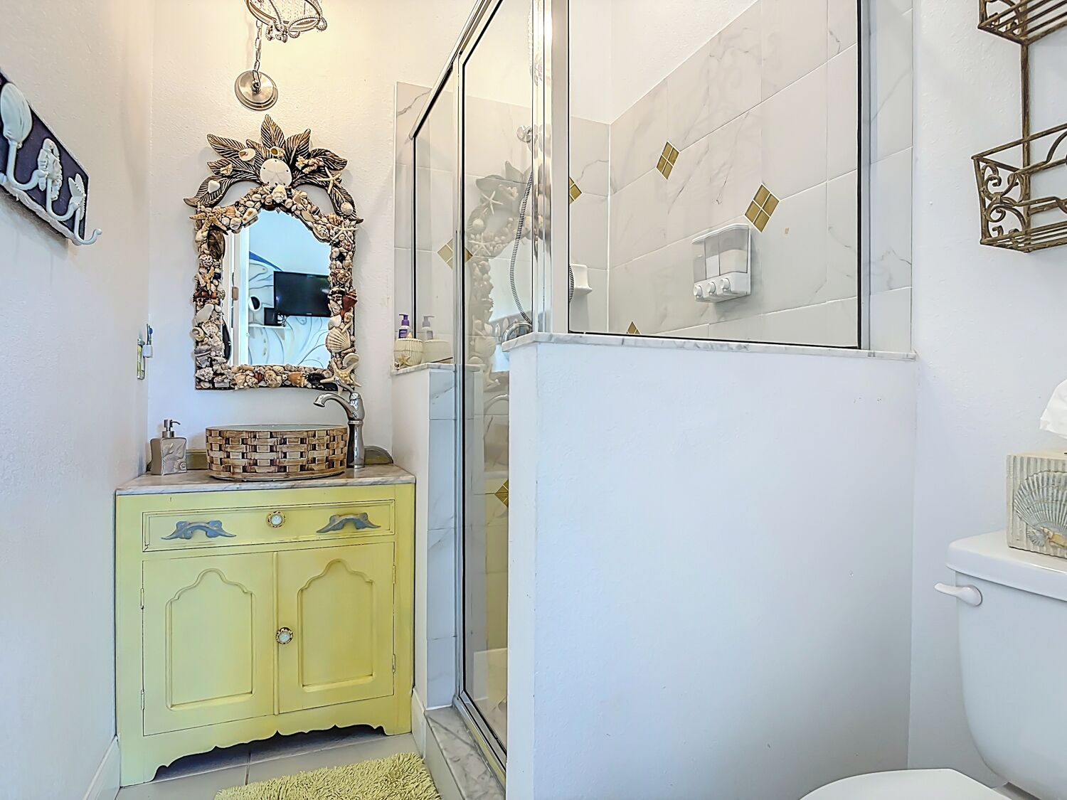 A bathroom with a yellow vanity, a large mirror with a decorative frame, a glass shower enclosure, and a toilet.