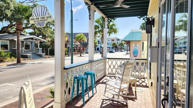 A covered outdoor seating area with a table and chairs, a sign for 'Inn on the Ave Bed and Breakfast', and a view of the street with buildings and palm trees. - Inn On the Avenue (New Smyrna Beach)