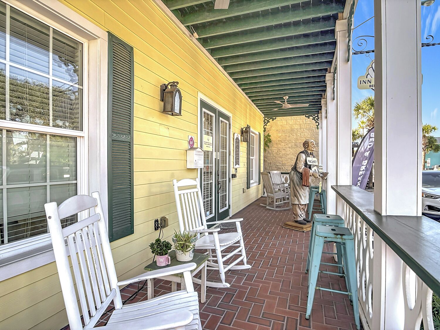 A yellow building with a covered patio area featuring white rocking chairs, a small table with potted plants, and a statue of a man holding a sign.