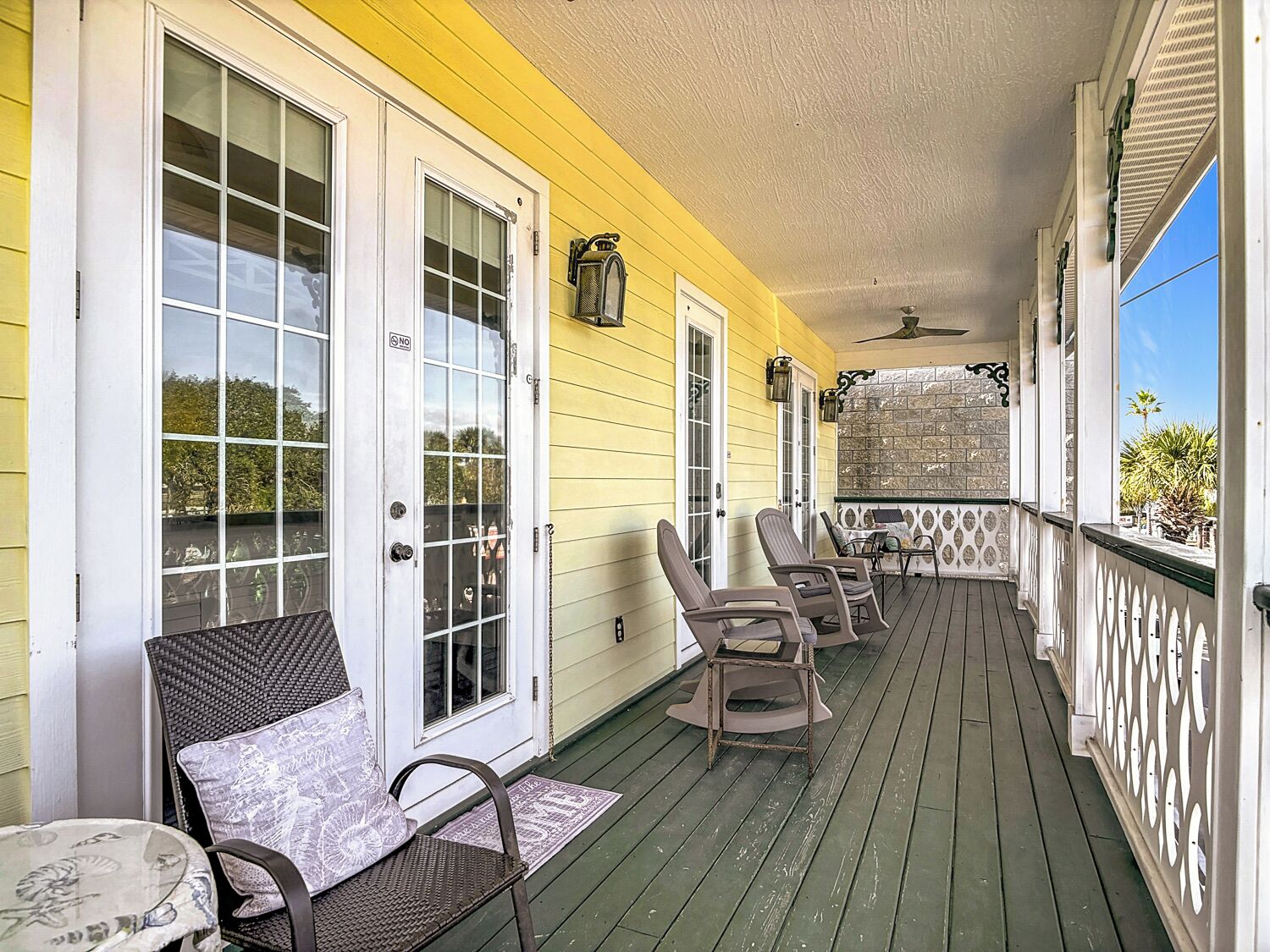 A covered porch with a green floor, white railings, and a stone wall. There are two chairs and a small table.
