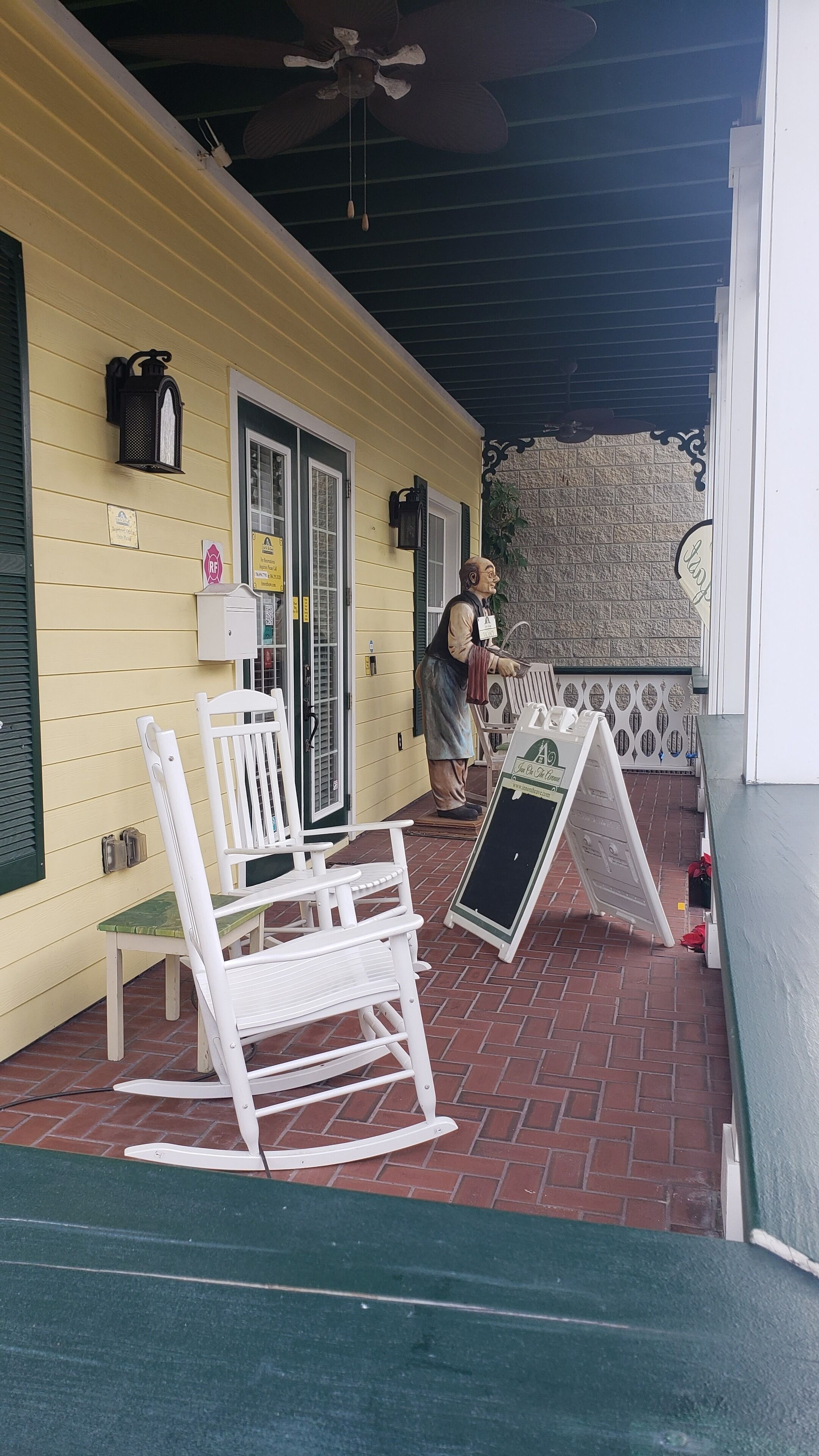 A yellow building with a covered porch, white rocking chairs, a statue, and a chalkboard sign.