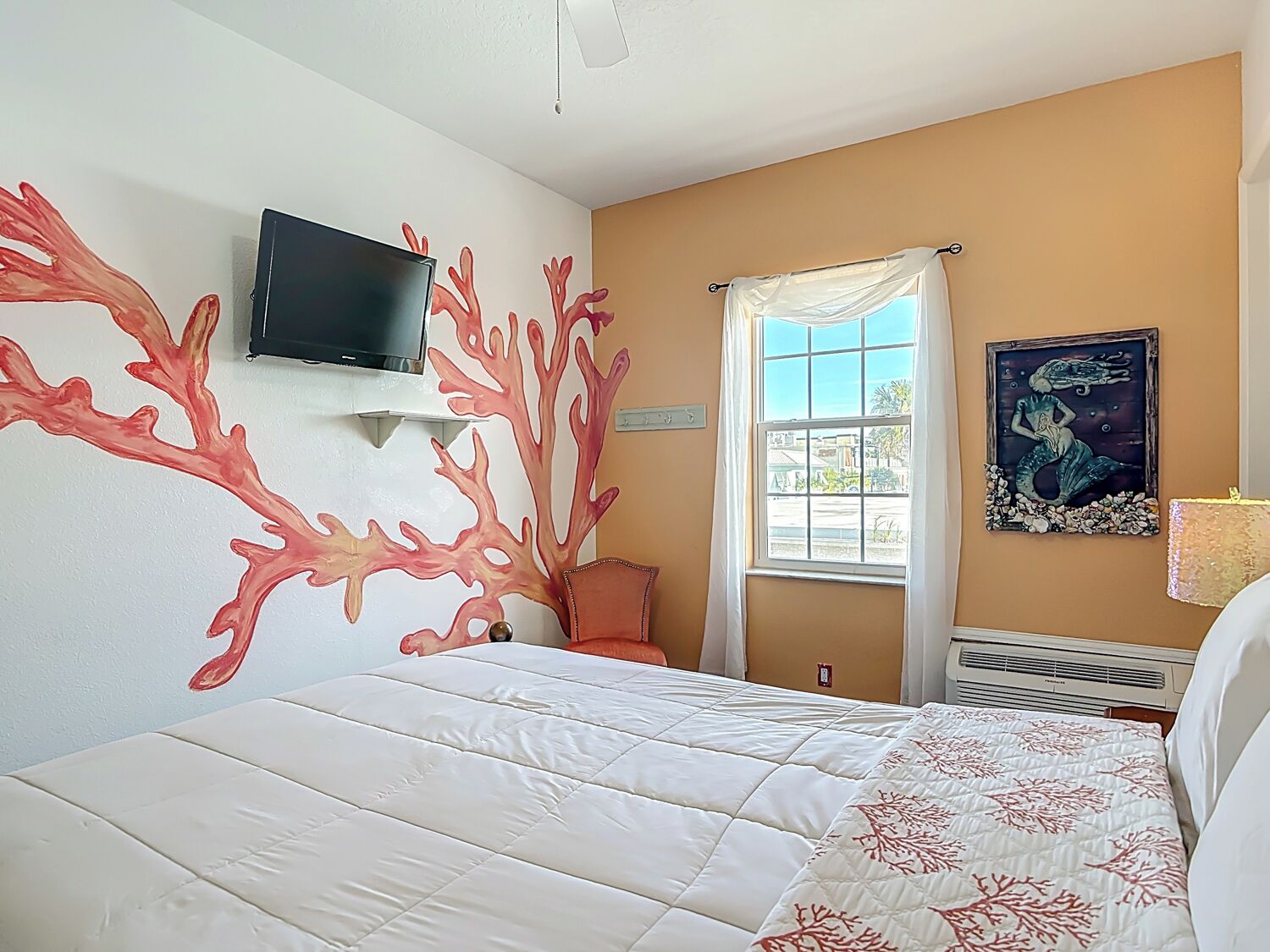 Habitación de hotel con cama, televisión montada en la pared, ventana con cortinas y un cuadro en la pared.