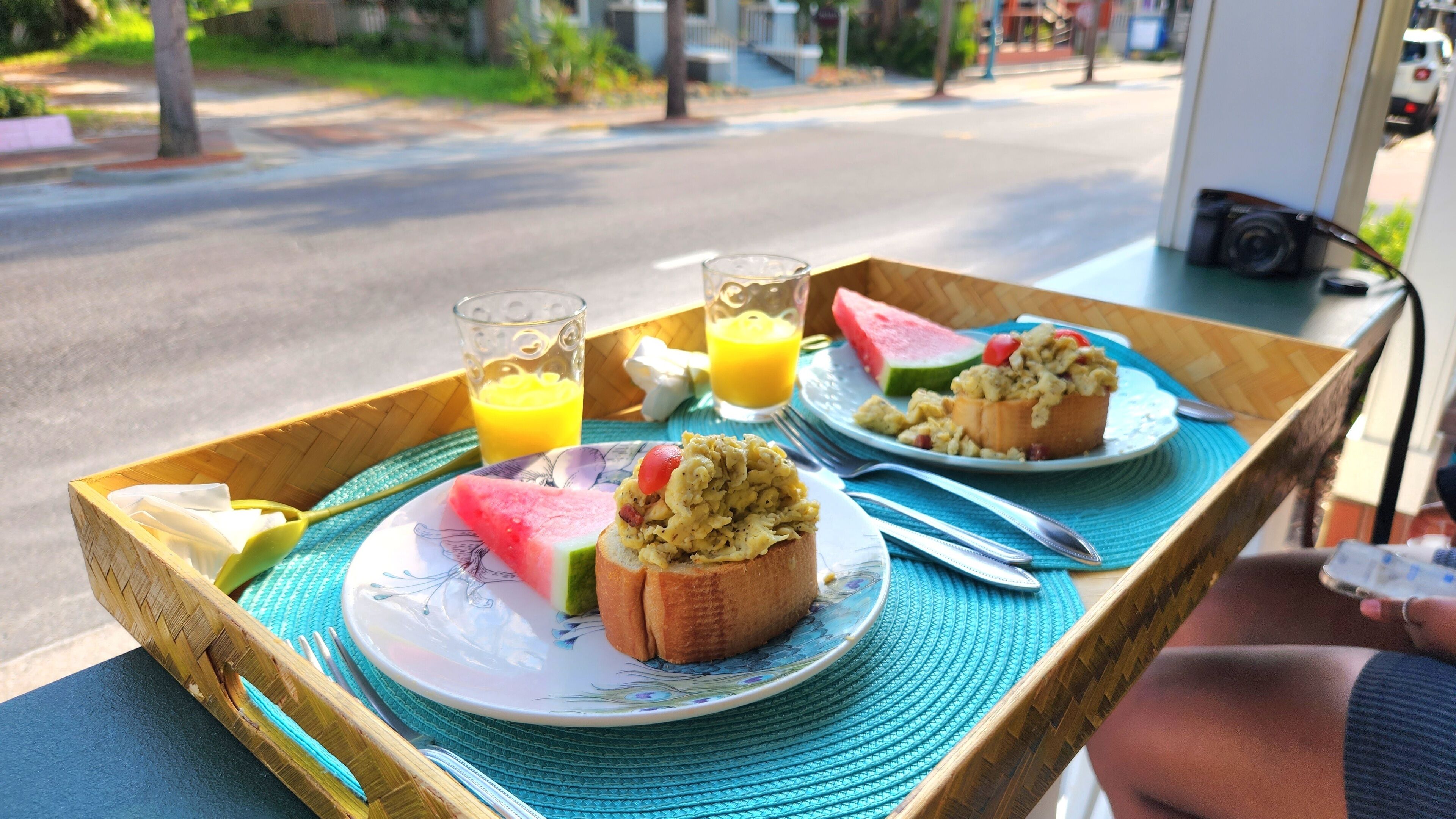 Aamiaistarjotin, jossa on paahtoleipää, munakokkelia, viipale vesimelonia ja kaksi lasillista appelsiinimehua, ja kaikki on asetettu punotulle alustalle.