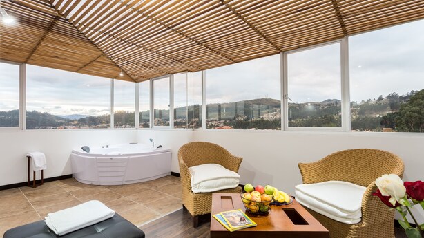 Una habitación moderna con un ventanal grande, un jacuzzi, mobiliario de mimbre y una mesa con frutas y una revista.