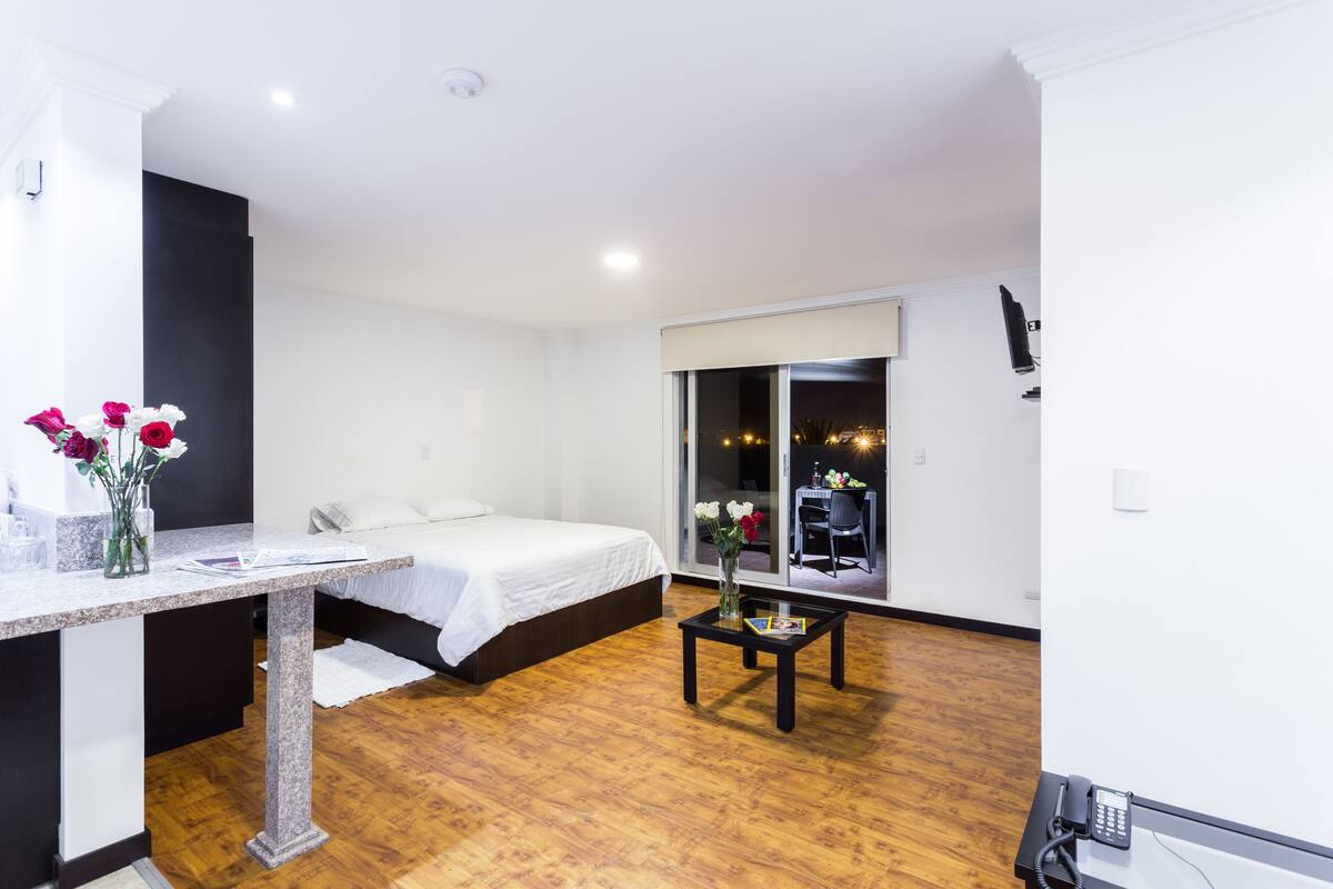 Habitación de hotel moderna con una cama, una mesita con un florero, un escritorio con un teléfono y vistas a la ciudad a través de amplias ventanas.