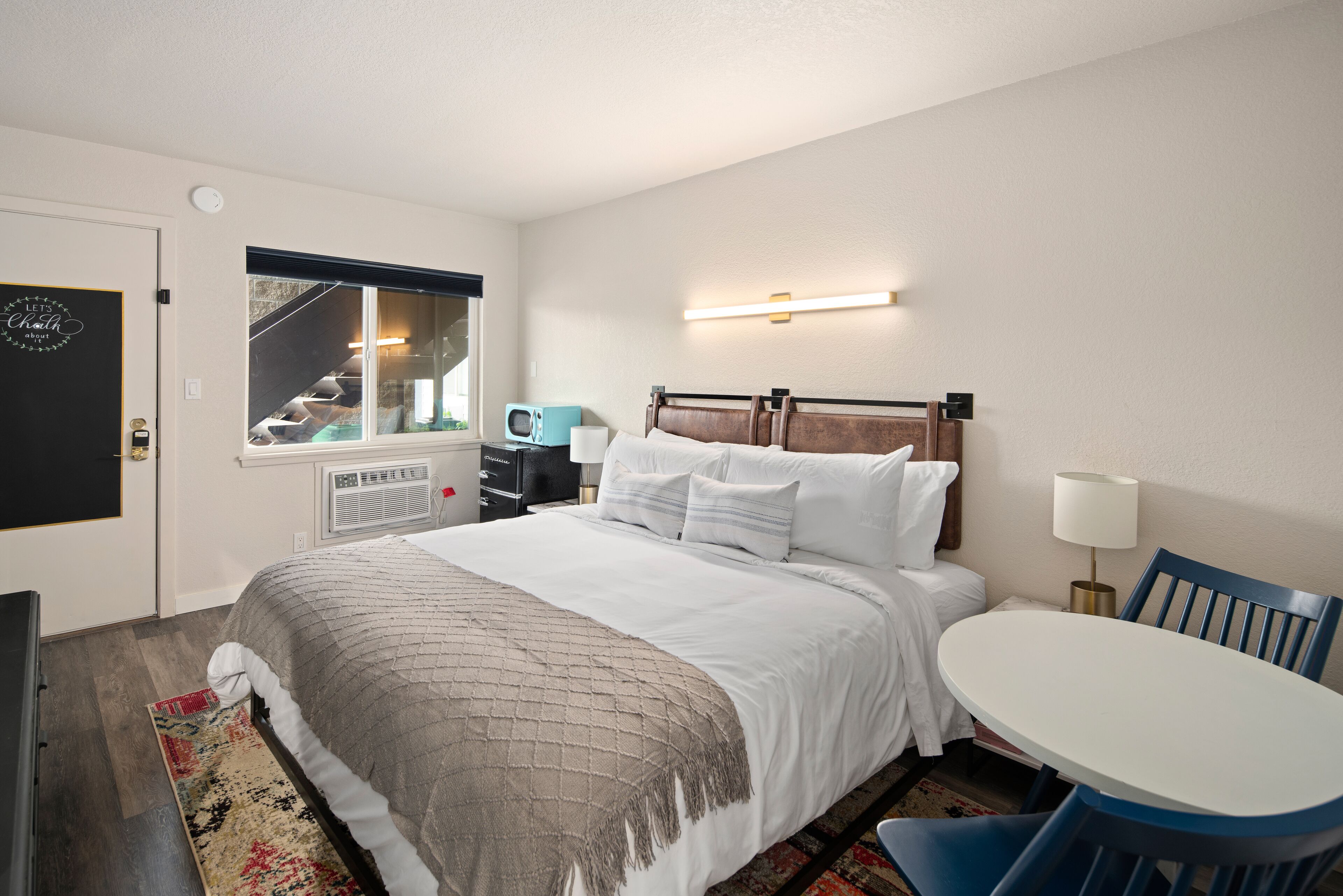 A hotel room with a bed, a desk, a chair, a window, and a wall-mounted light.