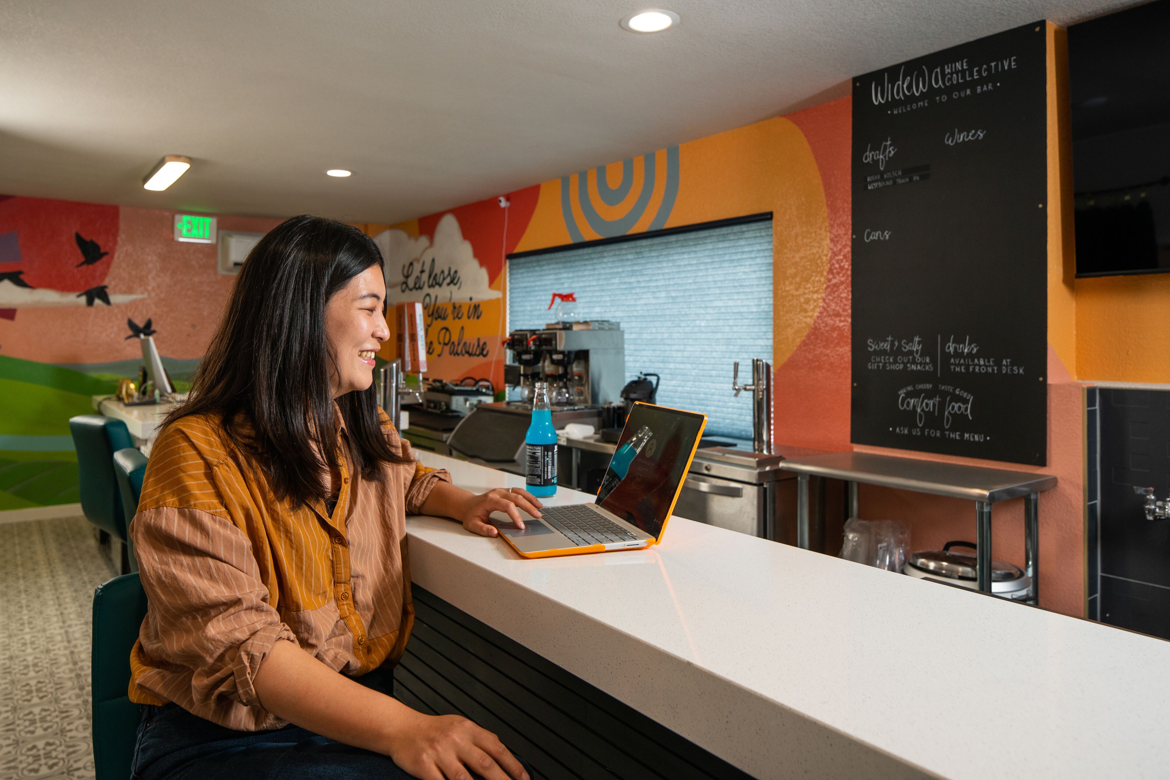 A woman working on a laptop at a counter with a menu board in the background.