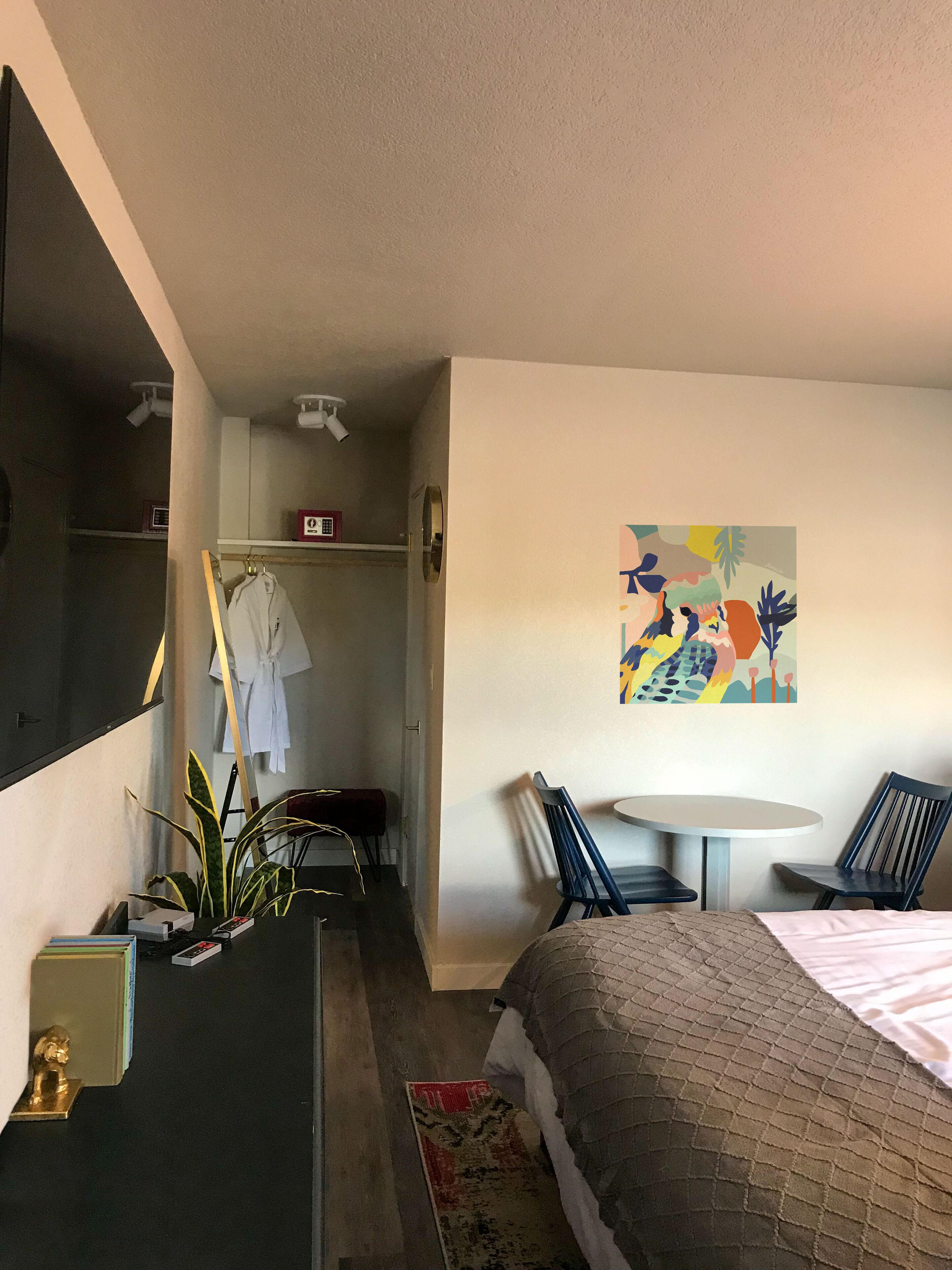 A hotel room with a bed, a desk, a chair, a small table, a plant, and a colorful abstract painting on the wall.