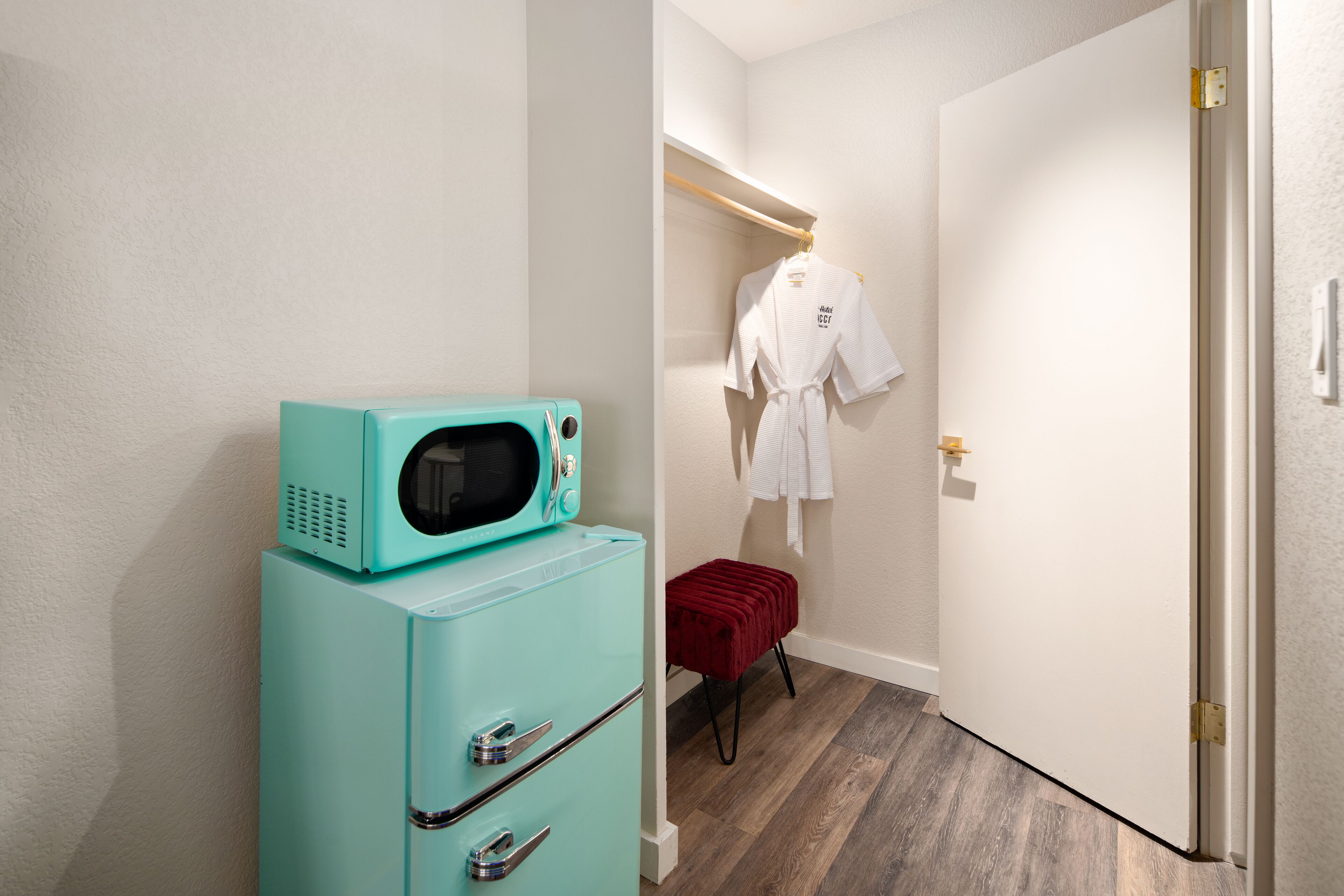 Habitación compacta con un microondas azul verdoso sobre un mueble, un taburete rojo y una bata blanca colgada en un perchero.