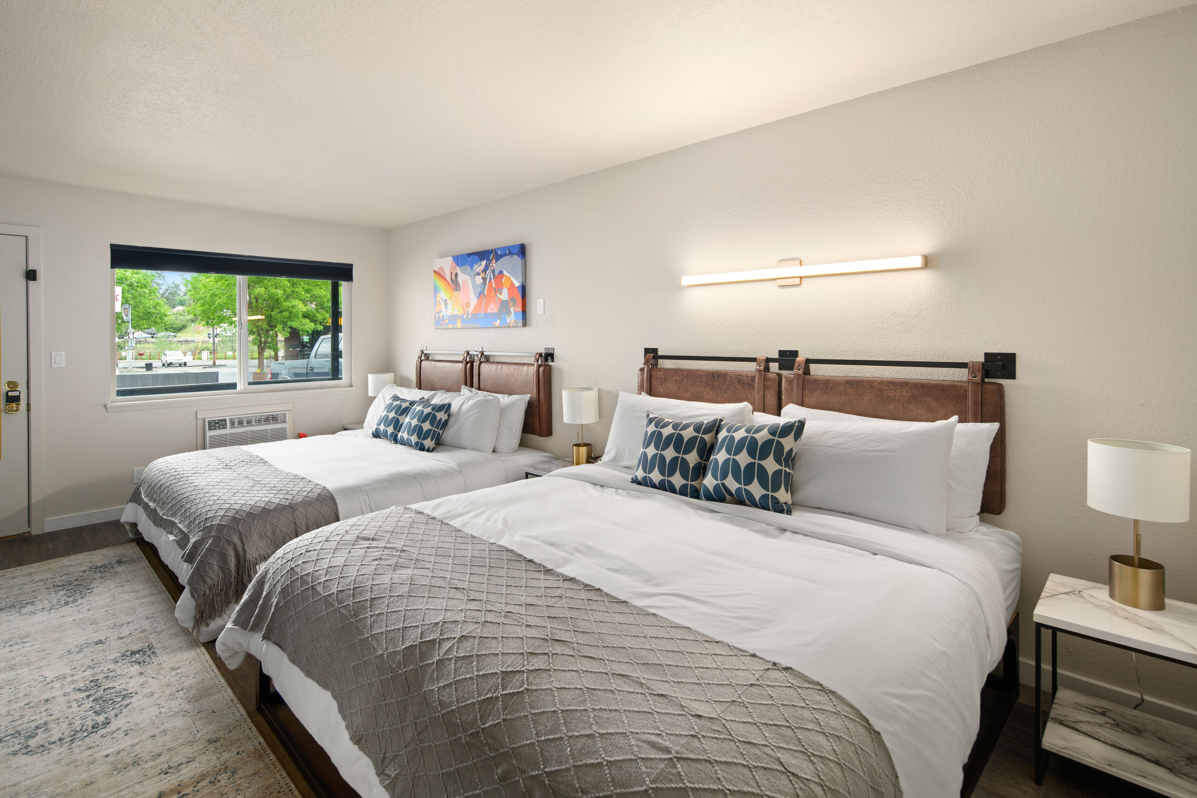 A hotel room with two beds, a large window, a painting, and a bedside table with a lamp.