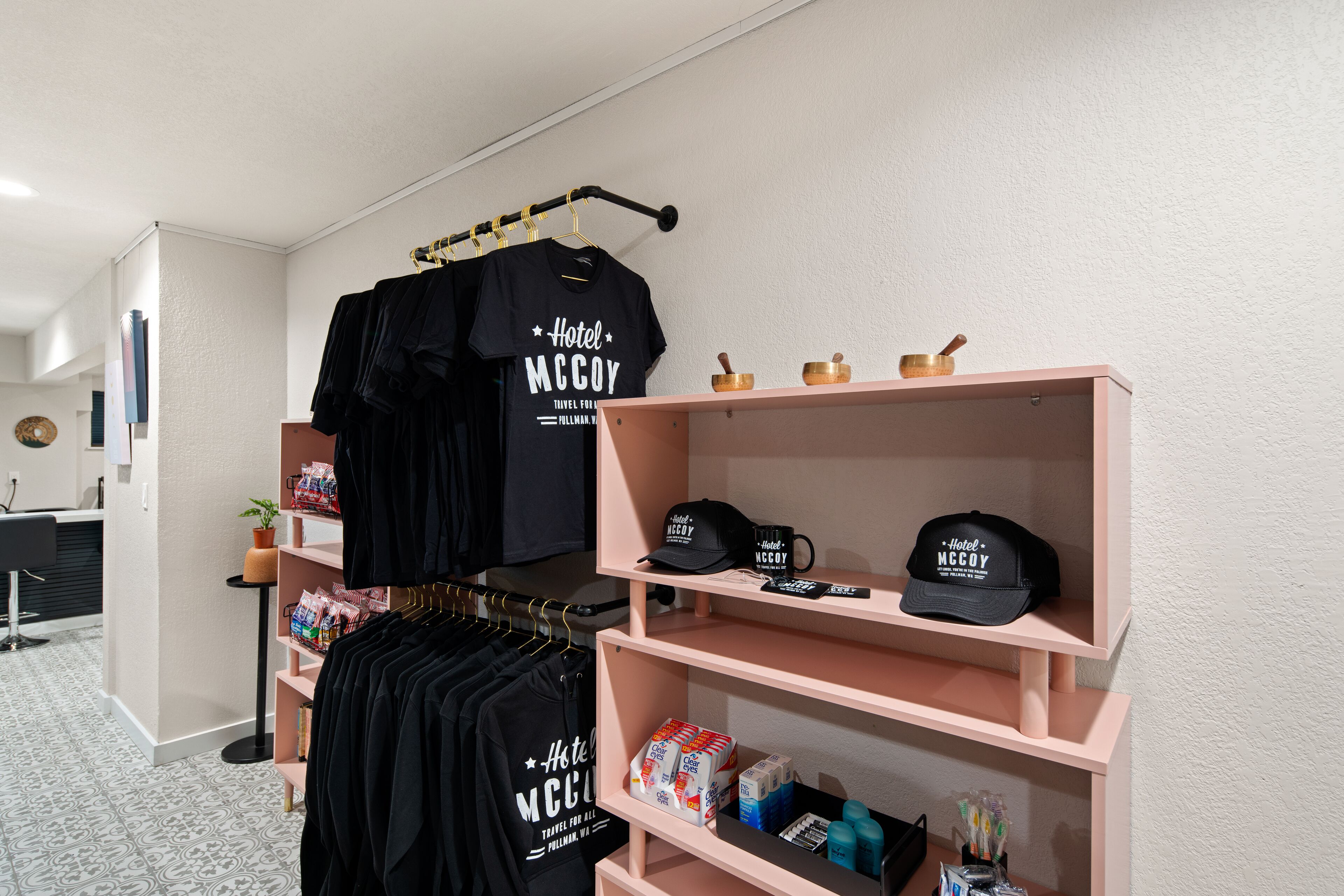 A hotel room with a bed, a desk, and a wardrobe. There are black t-shirts hanging on the wardrobe and a shelf with toiletries.