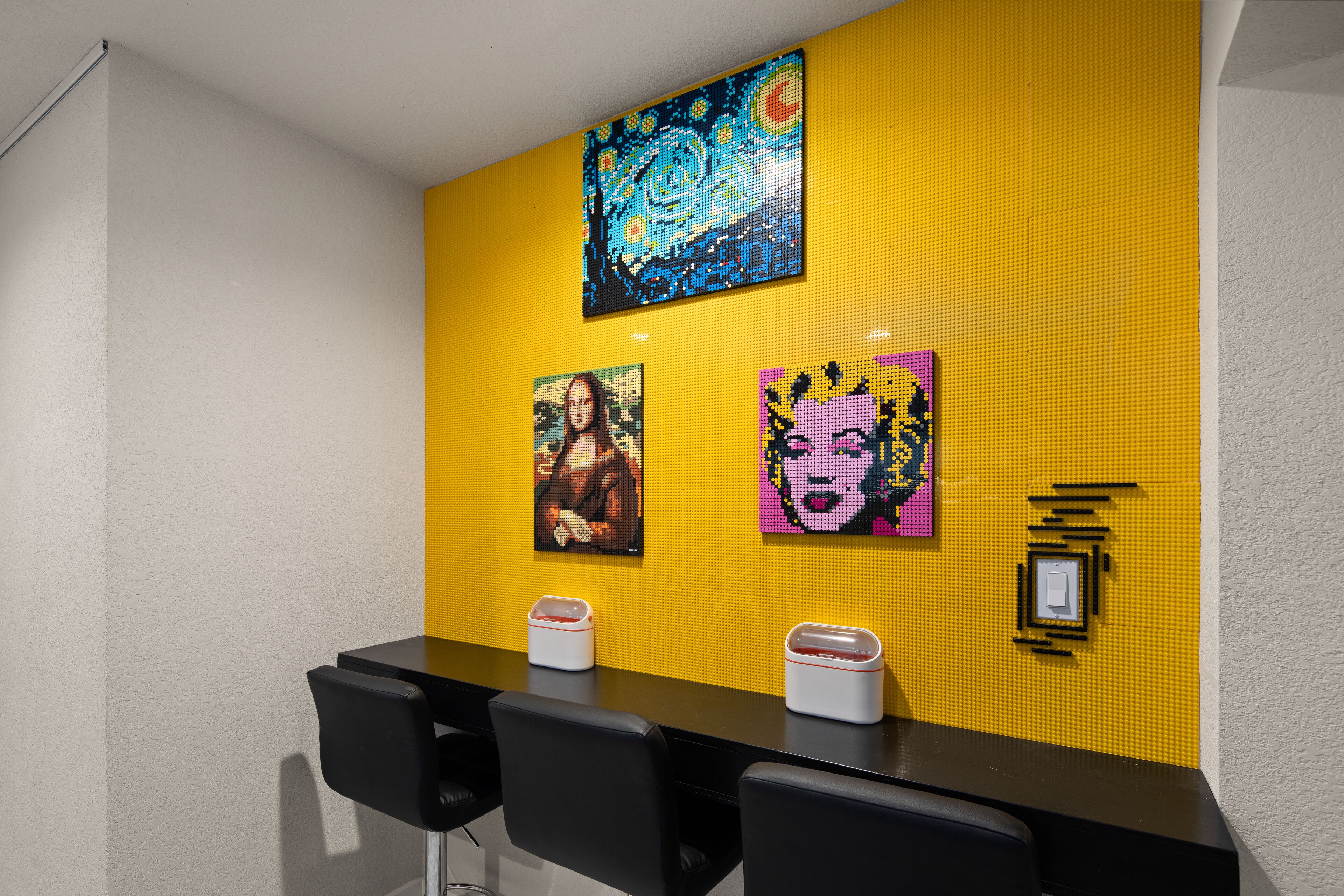 Una habitación con pared amarilla que presenta arte de píxeles de obras de arte famosas y una mesa negra con sillas.