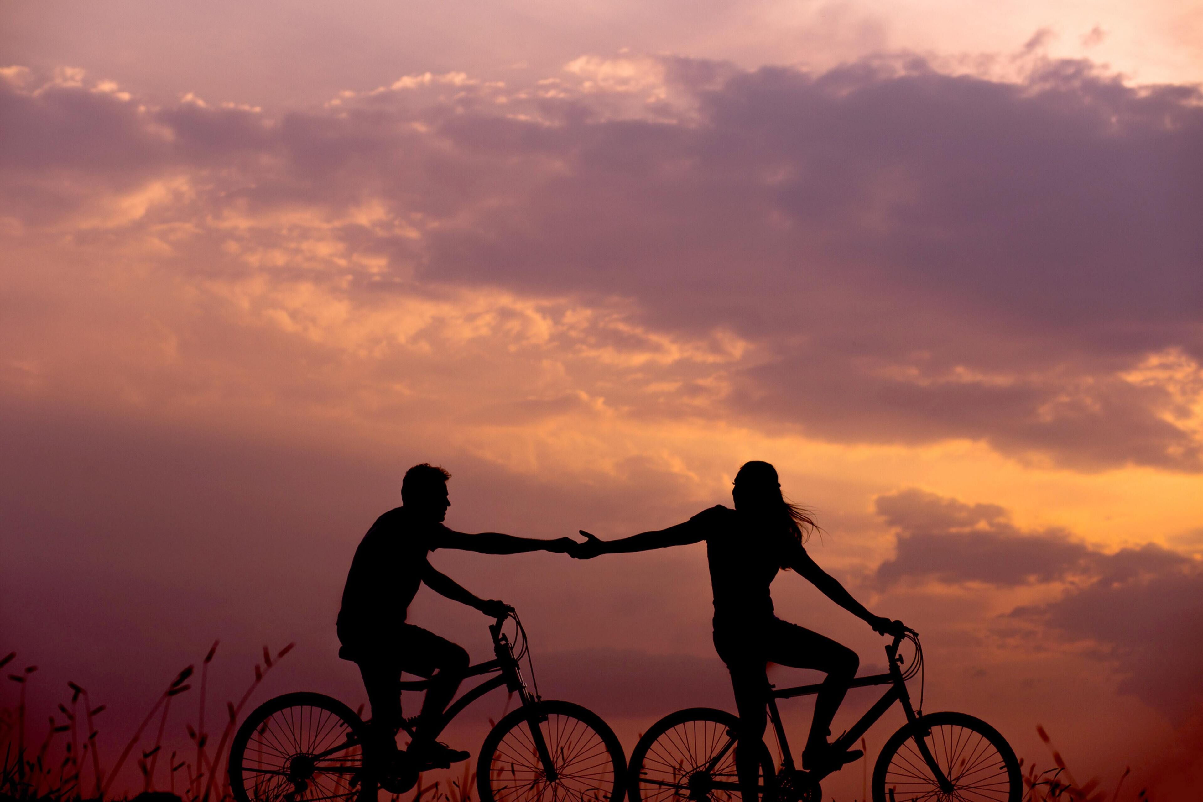 Two silhouetted figures holding hands on bicycles against a sunset sky.