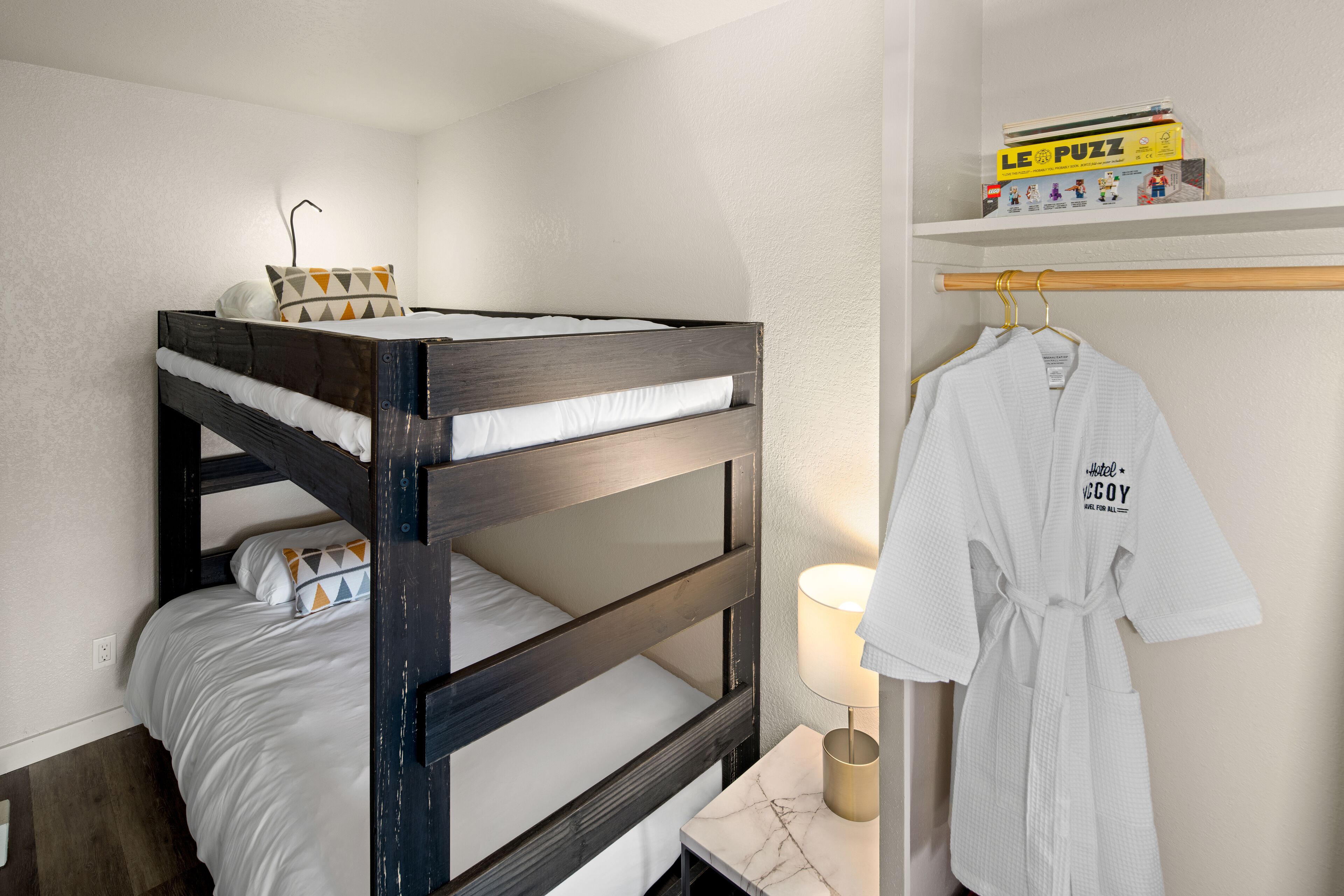 A bunk bed with white bedding, a bedside lamp, a robe hanging on a hook, and a shelf with a 'Le Puzz' box.