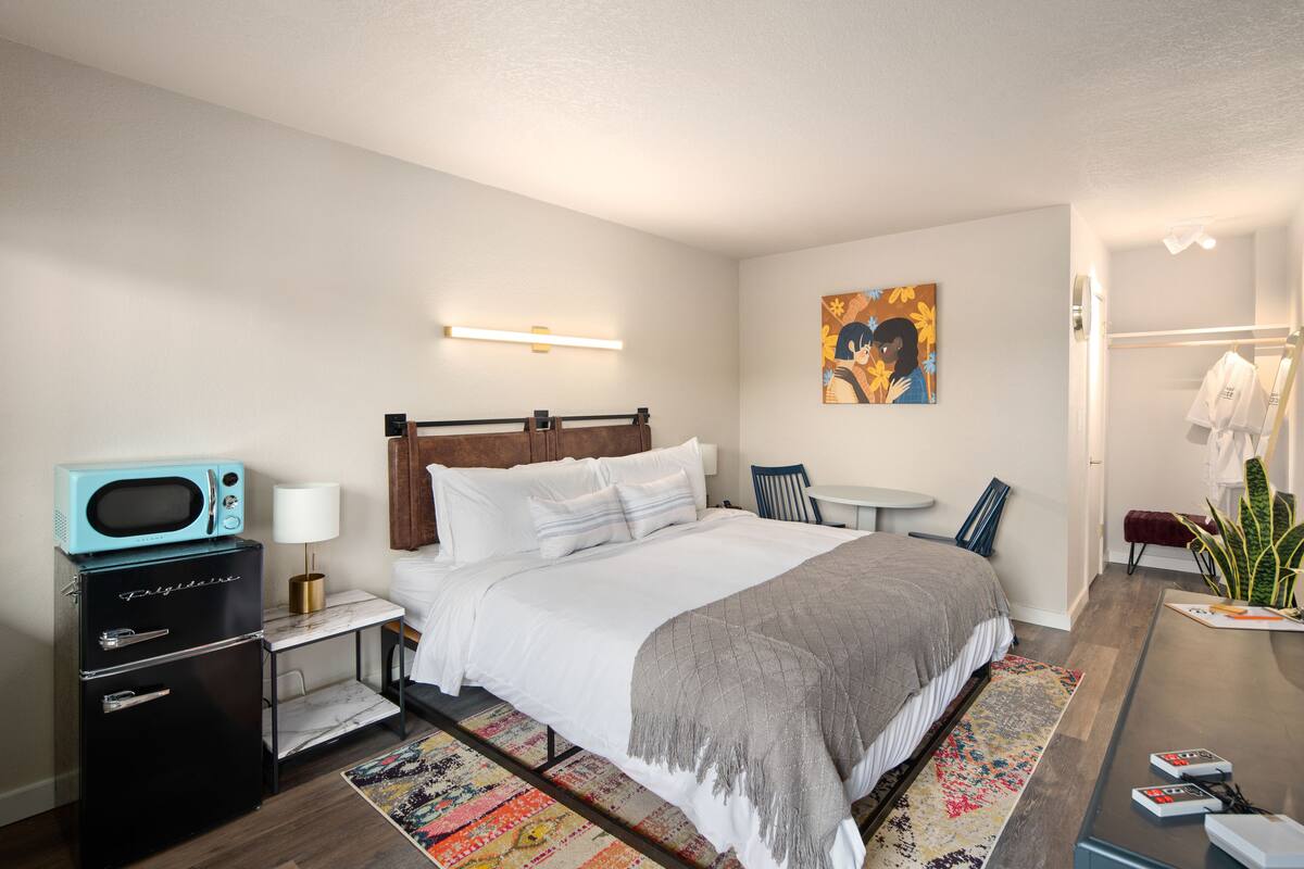 A modern hotel room with a bed, a small kitchenette with a microwave, a bedside table with a lamp, a chair, and a small table with a plant.