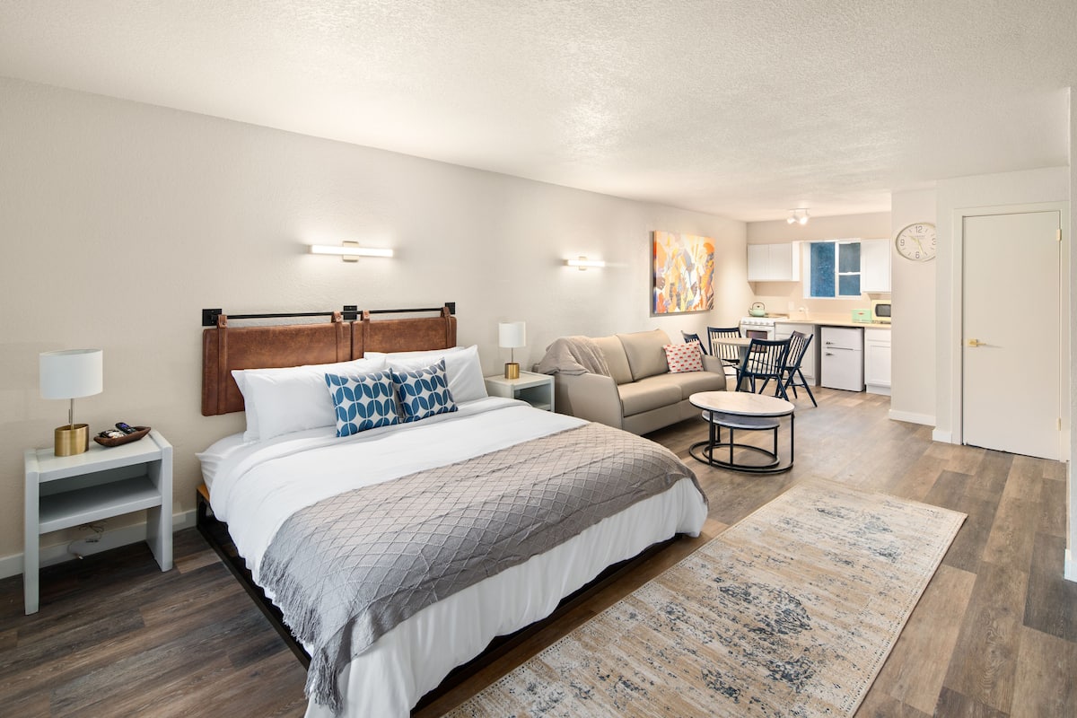 A bedroom with a bed, bedside tables, a nightstand, a sofa, a small round table, and a kitchen area in the background.