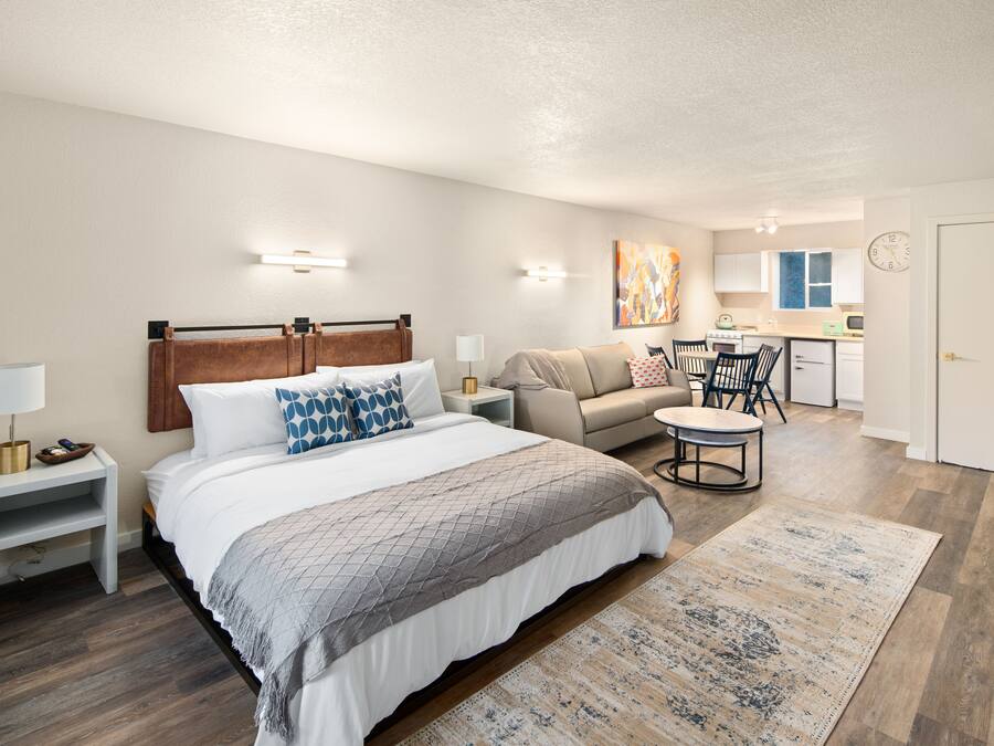 A bedroom with a bed, bedside tables, a nightstand, a sofa, a small round table, and a kitchen area in the background.