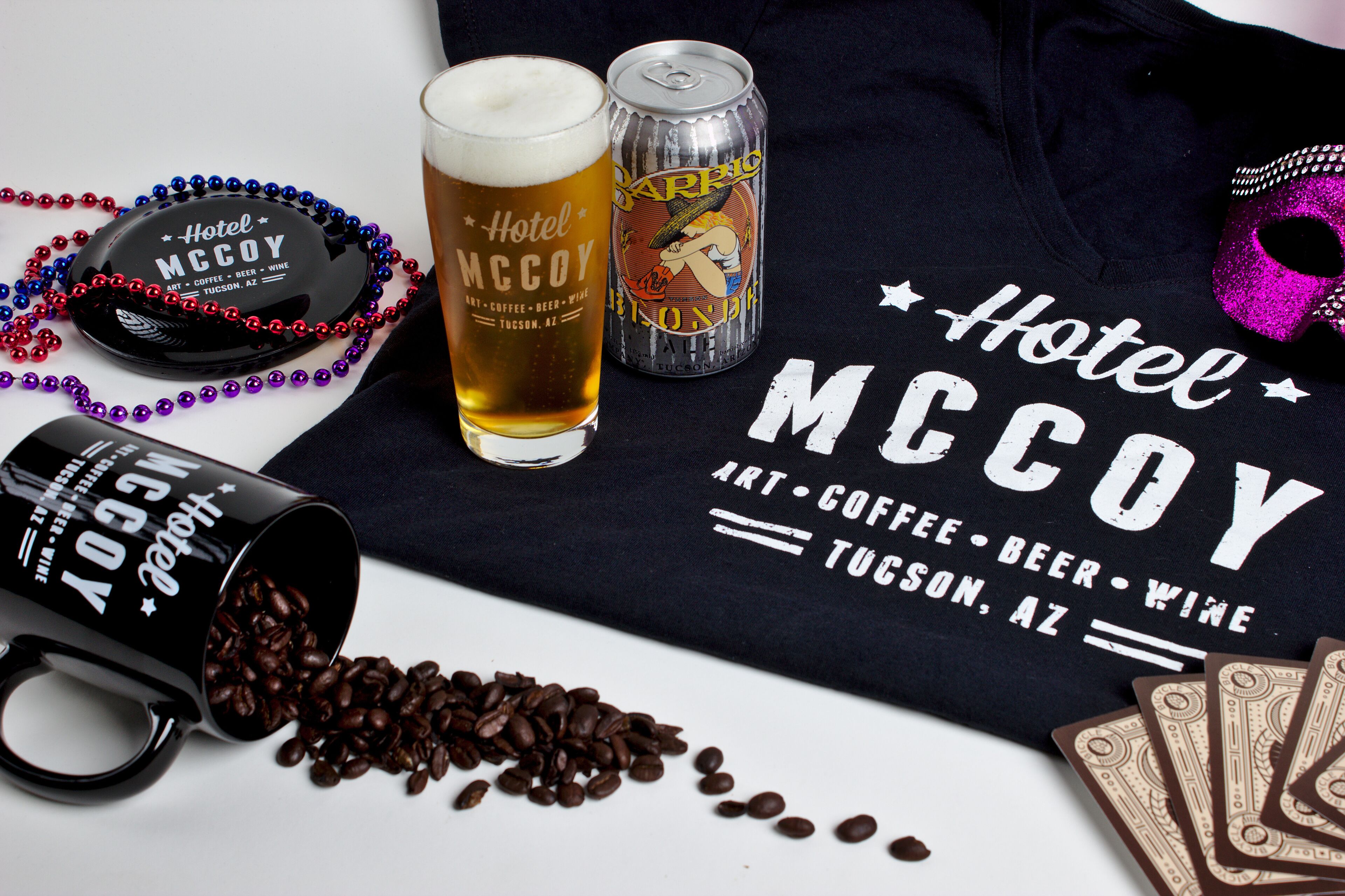A black mug with coffee beans spilled out, a glass of beer, a can of beer, and a black tray with a hat and beads.