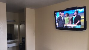A hotel room with a television mounted on the wall displaying a news program, a small wooden dresser, and a bathroom visible through an open door. - Bayview Motel (Emeryville)