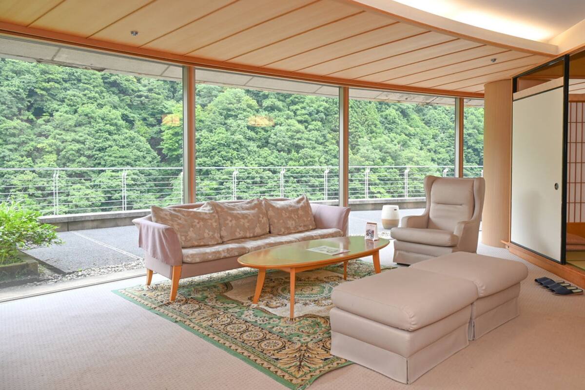 Una sala de estar moderna con sofá, sillón, reposapiés y mesa de centro. Amplias ventanas ofrecen vistas a un área boscosa.