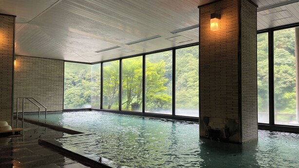 Une piscine intérieure avec des parois en verre transparent, une colonne en briques et une vue sur une zone boisée.