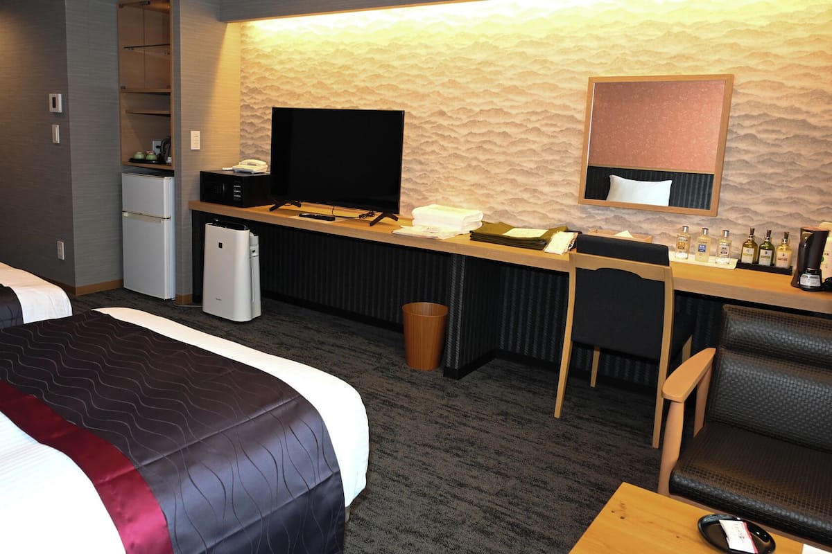 Habitación de hotel con dos camas, un escritorio con televisor de pantalla plana, un refrigerador, un cesto de basura y una zona de estar con una silla y una mesa.