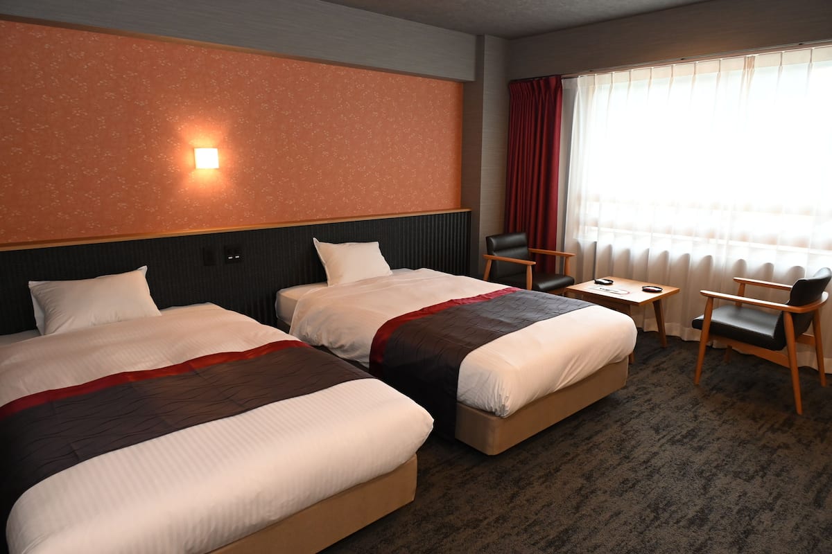 Habitación de hotel con dos camas, un escritorio, una silla y una ventana con cortinas.