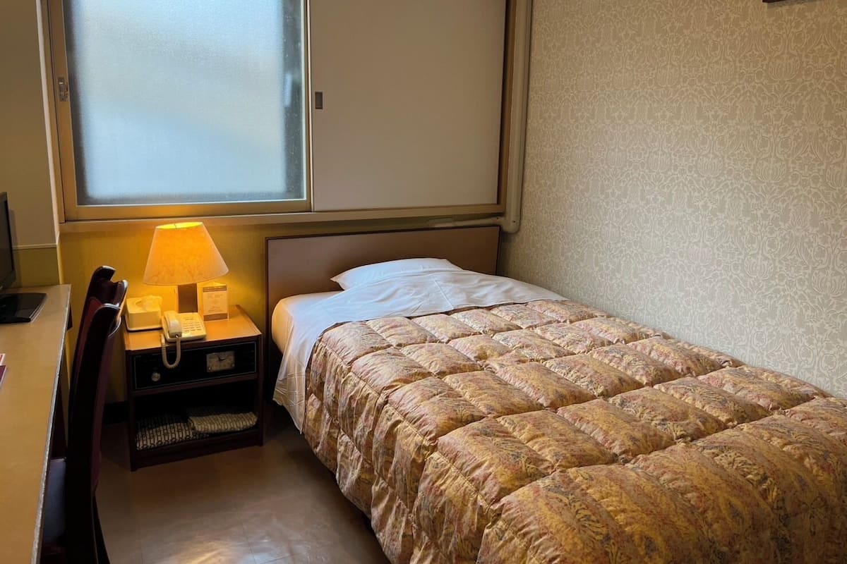 Quarto de hotel pequeno com uma cama de solteiro, uma mesa com abajur, telefone e janela com vidro fosco.